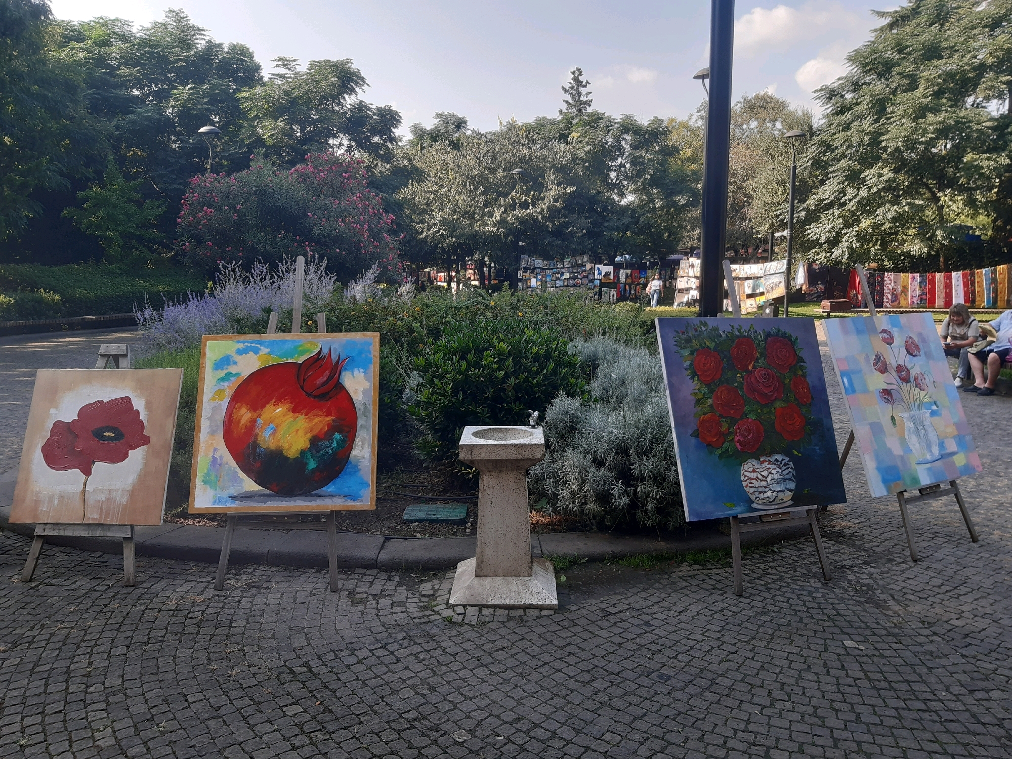 In the city of Tbilisi, Georgia, an outdoor art exhibition is taking place on a cobblestone path lined with lush greenery. Four paintings are displayed on easels in the foreground. The first painting to the left depicts a red poppy on a beige background. Next to it, a vibrant painting of a red apple with abstract colors in the background is showcased. To the right, a still life of red roses in a decorative vase is presented, followed by a painting of tulips in a glass vase against a pastel-colored backdrop. In the background, several people are browsing through other artworks displayed on racks and tables, suggesting a vibrant marketplace atmosphere. Trees and flowering bushes form a natural backdrop, with the sky visible overhead. A stone water fountain stands between the paintings, adding to the ambiance of the outdoor setting.