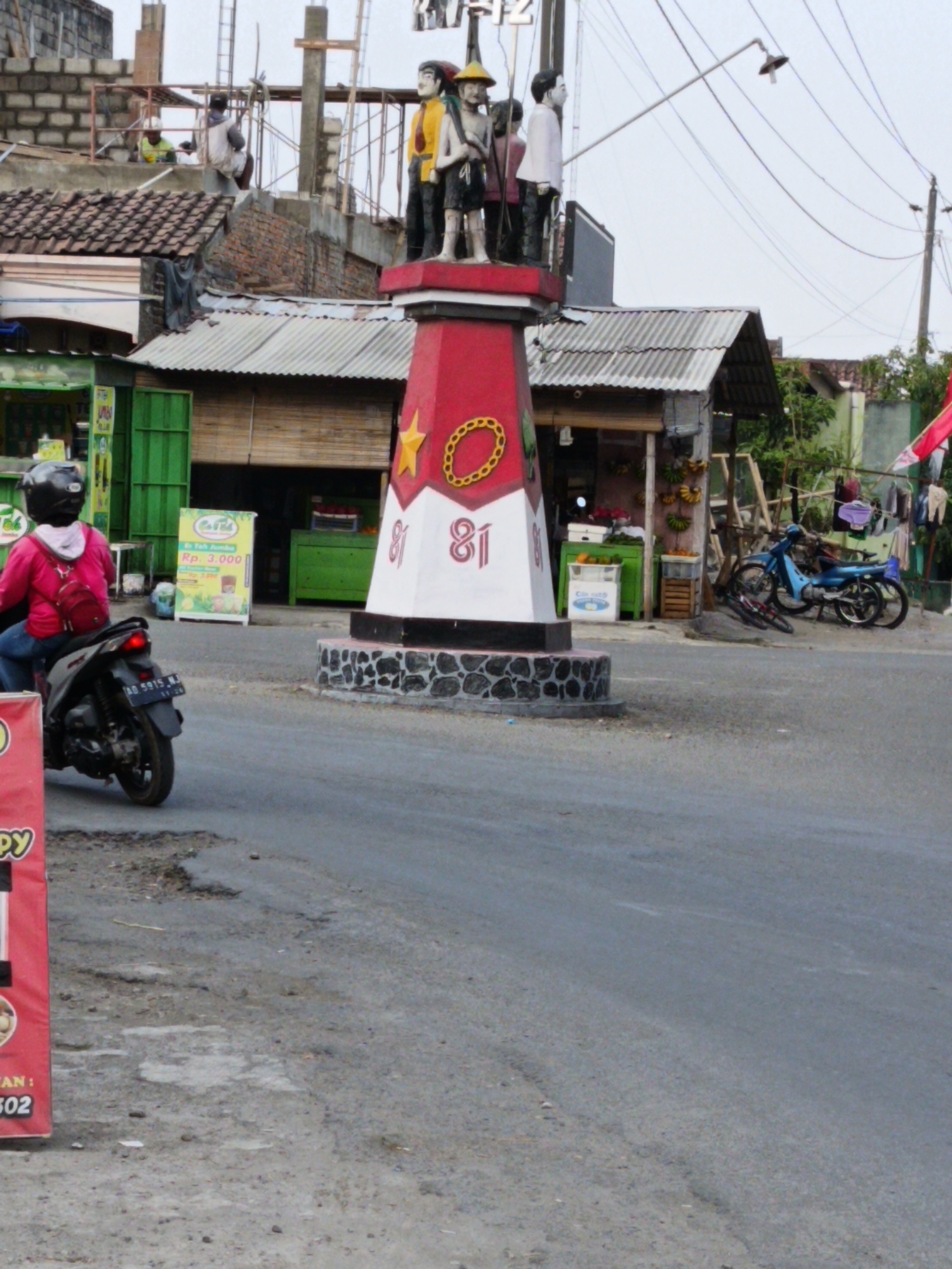 The image shows a street scene in Sukorini, Indonesia. In the foreground, a person wearing a pink jacket and a black helmet is riding a scooter. The scooter has a license plate that reads "AD 5915 MJ". The street is paved but appears to have some potholes.
In the background, there is a monument with several statues of people. The monument is red and white and has the number "81" on it. Next to the monument are small shops with corrugated metal roofs. One shop has a green facade and displays fruit such as bananas and oranges. There is also a blue motorcycle parked on the side of the road. Clothes are hanging on a line to dry in front of one of the shops. The sky is overcast.
The overall scene depicts a typical street in a developing urban or semi-urban area, with a mix of residential and commercial buildings, various modes of transportation, and everyday activities taking place.