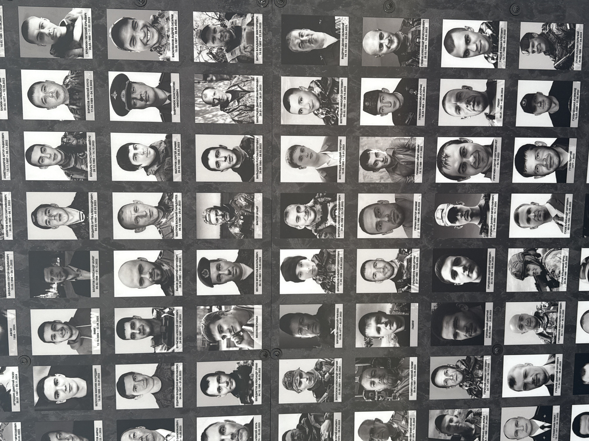The image shows a memorial wall in Moscow, Russia. The wall consists of numerous black and white portrait photos arranged in rows and columns. Each photo is accompanied by text, likely names and dates. The people in the portraits appear to be soldiers, some in uniform, others in civilian clothing. The overall impression is that of a memorial to fallen soldiers.