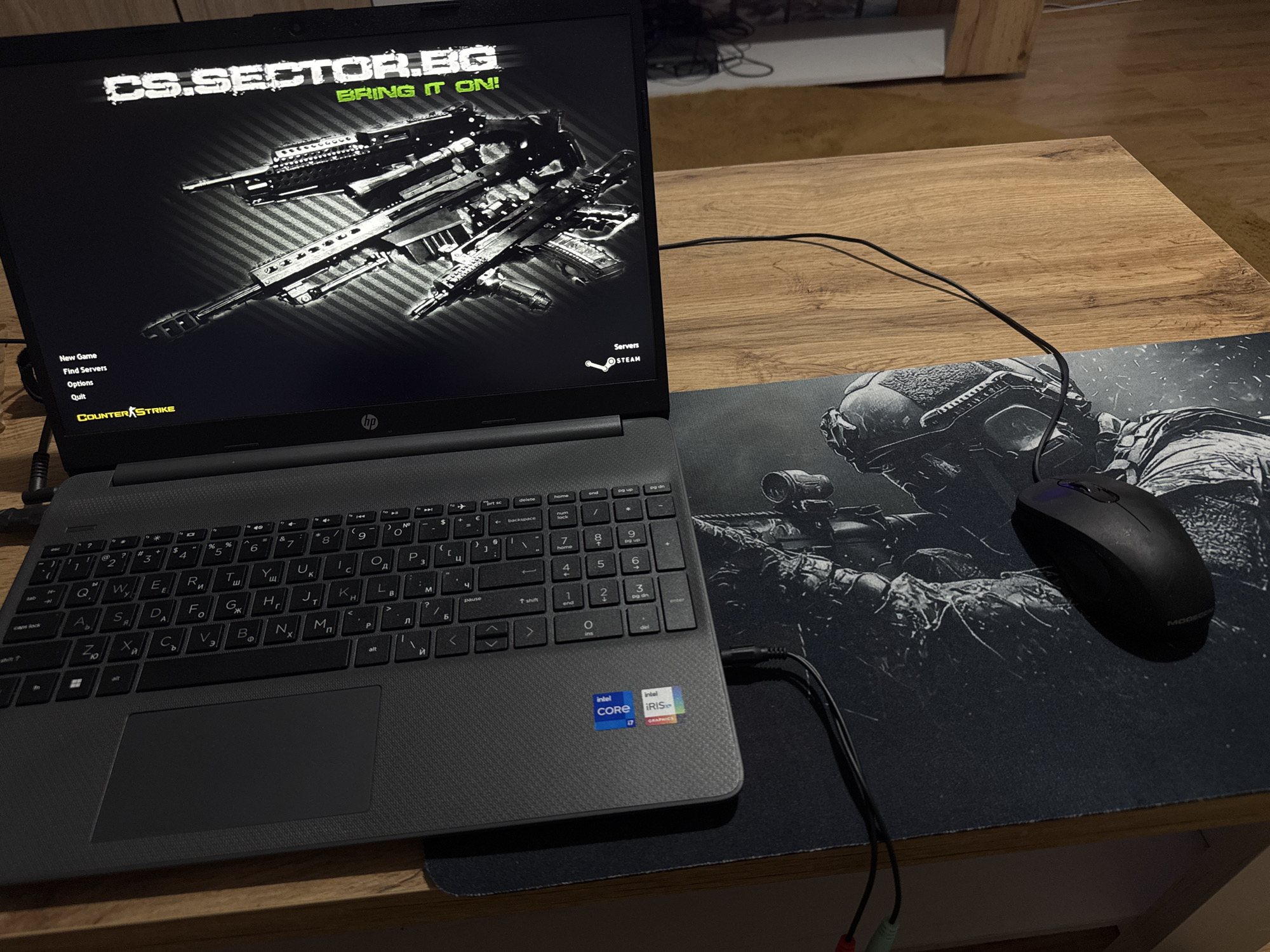 A black HP laptop is open on a wooden desk, displaying the Counter-Strike game menu. The screen shows the game's logo "CS.SECTOR.BG BRING IT ON!" and two assault rifles. On the left side of the screen are menu options: "New Game," "Find Servers," "Options," and "Quit." Below these options is the Counter-Strike logo. To the right of the game menu, the "STEAM" logo is visible. The laptop's keyboard is black with Cyrillic characters. Below the keyboard is a large touchpad. To the right of the laptop, on a black mousepad featuring a soldier aiming a rifle, is a black computer mouse. The mousepad is large and covers a significant portion of the desk. The environment appears to be indoors, with wooden flooring and furniture visible in the background. The lighting is dim. The image does not provide any specific location information within the city of Ruse, Bulgaria. There are no people or animals present in the image.