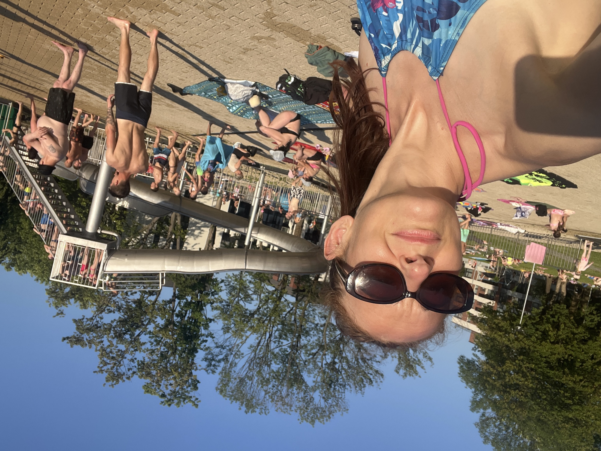 This media file shows a group of people at what appears to be a water park or swimming pool area on a sunny day. In the foreground, a woman is taking a selfie, wearing a blue and floral patterned bikini top and dark sunglasses. Her hair is dark brown and appears to be wet.
In the background, several other people are visible. Some are standing near a water slide structure, while others are lying on towels or walking around the paved area. There are also glimpses of trees and a blue sky, suggesting an outdoor setting. The overall impression is one of a relaxed, recreational atmosphere. The sunlight indicates it is daytime. There is no visible text in the image.