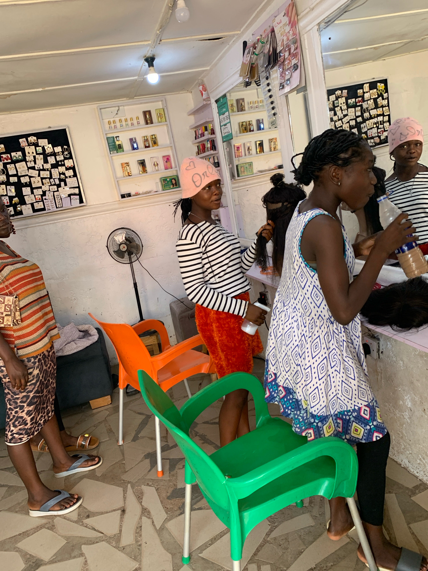 The image depicts a scene inside a hair salon located in Jos, Nigeria. Three young females are visible, along with various hair-related products and accessories displayed on the walls. One female, wearing a striped long-sleeved shirt and an orange skirt, is holding a spray bottle. Another female, in a white dress with a blue and white pattern, is holding a bottle of liquid and appears to be in the process of styling hair. The third female, wearing a pink head covering, is also engaged in hair-related activities. The salon environment includes display shelves with hair products, earrings on a board, a fan, and chairs. The floor is tiled with an irregular pattern.