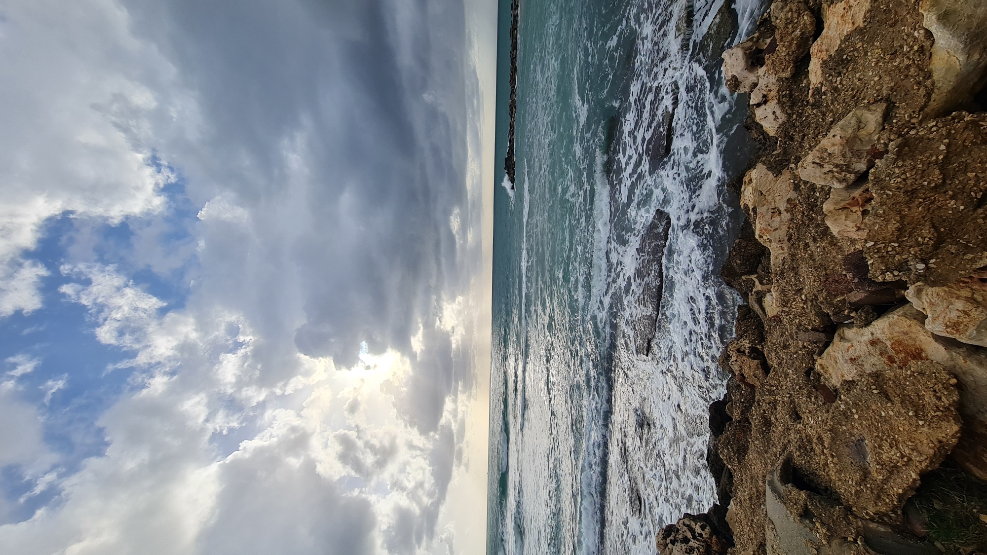 The image depicts a coastline in Nahariyya, Israel. The composition is divided vertically, with a cloudy sky on the left and the sea on the right. The sea is turbulent, with waves crashing against a rocky shoreline on the right. The sky shows a mix of dark and light clouds, and the sun is partially obscured. The photo suggests a potentially stormy or overcast day.
