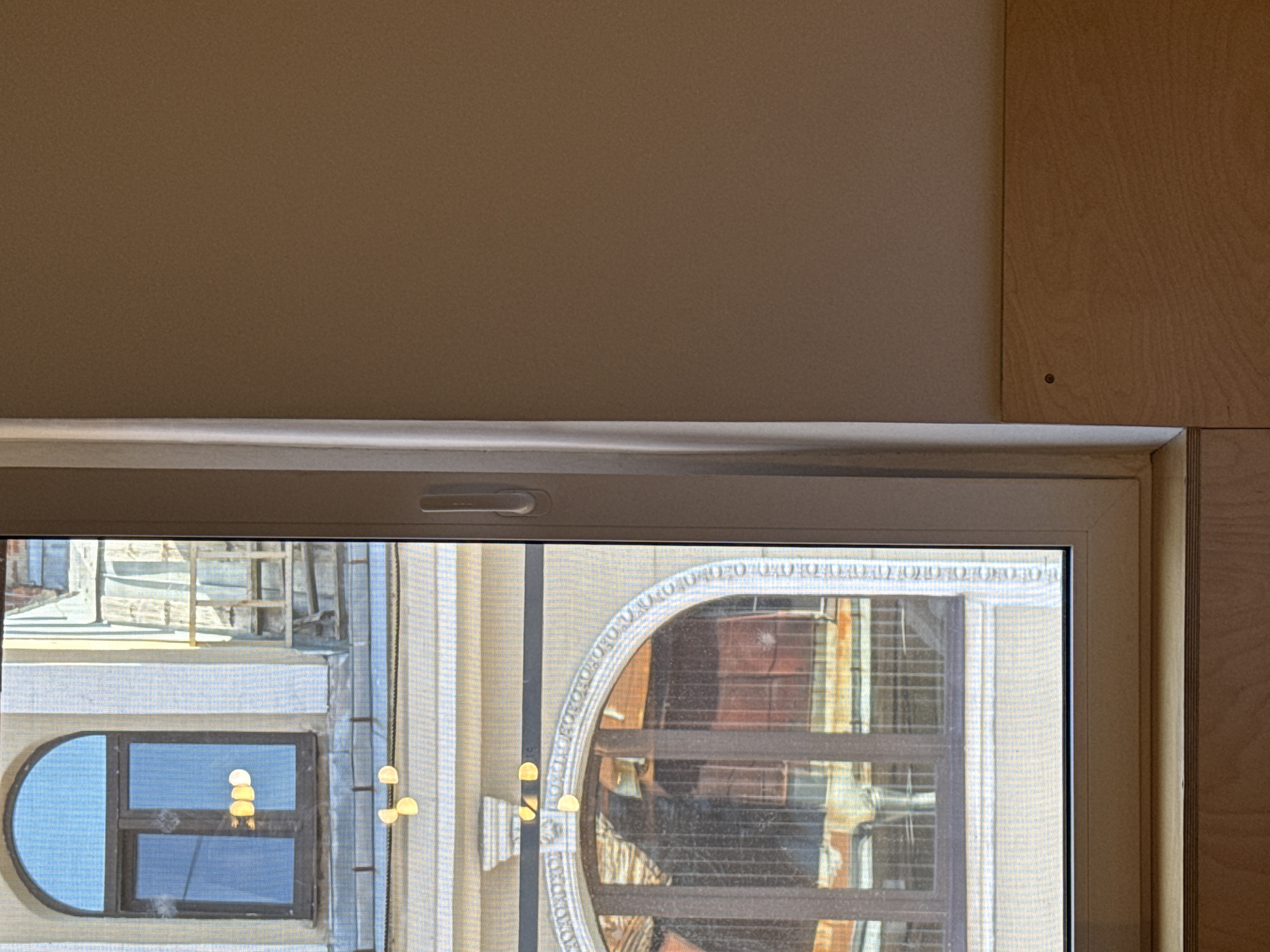 The image shows a close-up view of a modern window with a white frame, looking out onto an older building. The window has a screen, and through it, one can see arched windows and architectural details of the exterior building. There are some reflective, yellow circular objects visible both inside the window and on a string outside. The interior of the room consists of a plain, light-colored wall and a wooden surface below the window. The scene appears to be an interior space looking out onto a historical building, likely in Saint Petersburg, Russia, given the architectural style of the exterior.