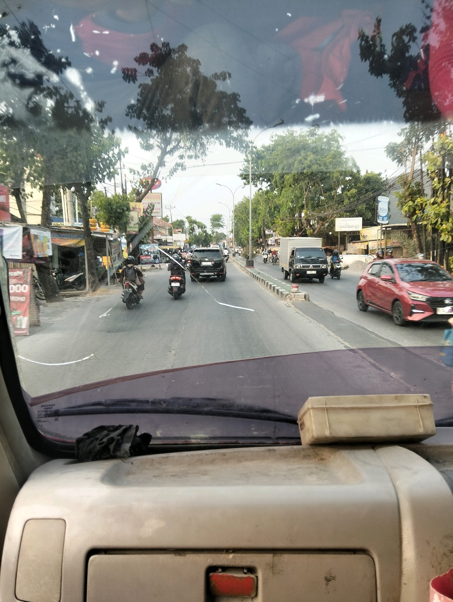 The image is taken from inside a vehicle, looking out through the windshield at a street scene in Semarang, Indonesia. The windshield is dirty and streaked, with reflections of trees and red fabric. In the foreground, on the dashboard, are a black cloth and a small, light-colored plastic box.
The street is paved with asphalt and appears to be a multi-lane road with a median strip. On either side of the road are buildings, trees, and sidewalks. There is moderate traffic, with several vehicles visible. A red car is in the right lane, a black SUV is further up on the left, and a white box truck is in the left lane. Several motorcycles are also present, with riders wearing helmets. Some people are visible on the sidewalks, and some are operating motorcycles.
There are signs visible on the buildings and along the roadside, though the text is mostly illegible. One sign on the left side of the road appears to be for a business with the word "000" in large red letters. Another sign further down the road has the word "TRA" visible. The overall impression is a bustling urban street scene during daylight hours. The presence of the reflective fabric on the dashboard suggests the vehicle may be parked or moving slowly, with the sun at an angle that causes reflections on the windshield.