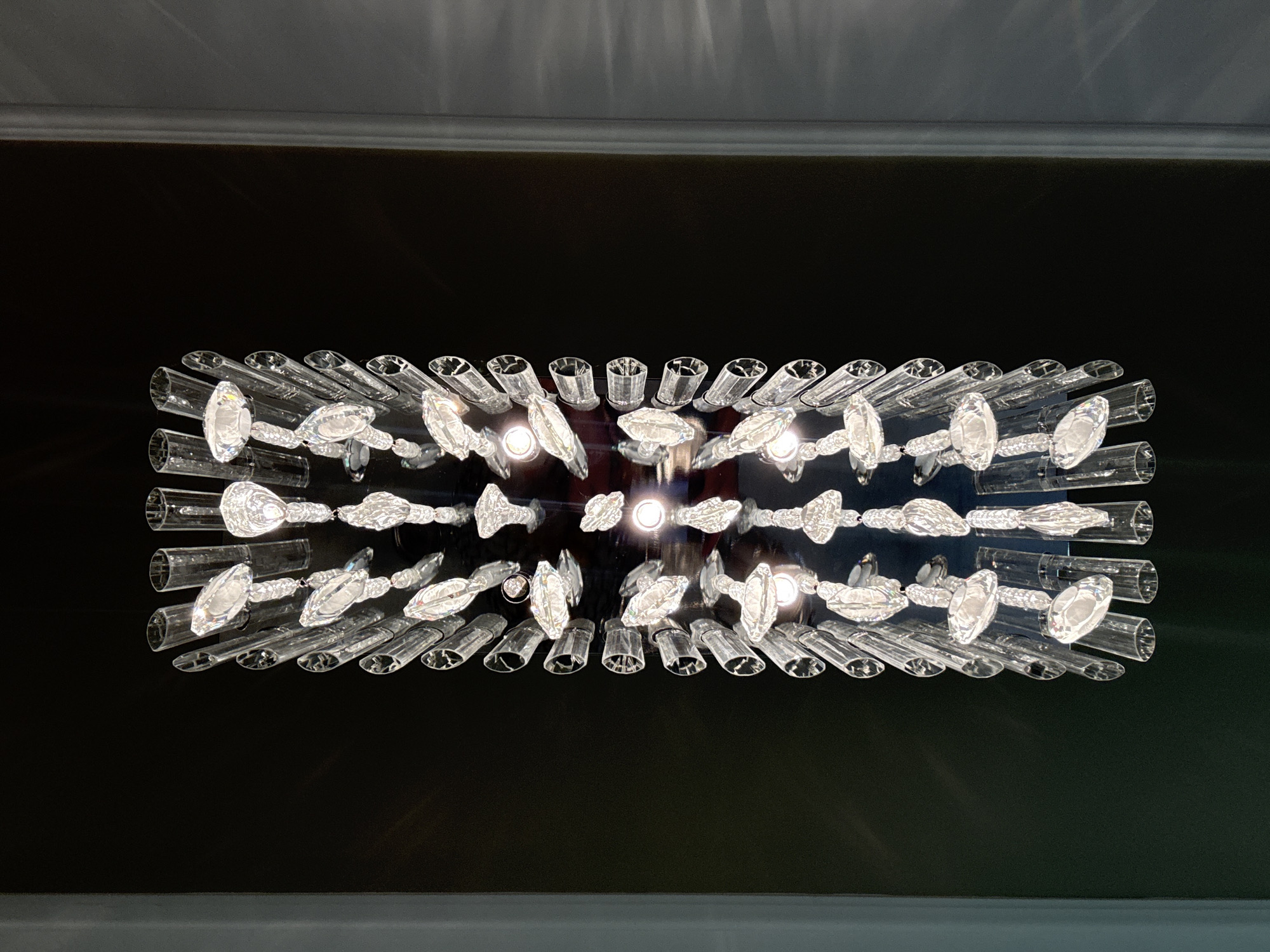The image shows a rectangular, modern chandelier mounted on a dark-colored wall, likely in an indoor setting. The chandelier features clear glass tubes arranged in rows, with crystal-like elements attached between the tubes. Light bulbs are visible in the center of the chandelier, providing illumination. The wall is dark, possibly black or a very dark shade of gray, contrasting with the bright chandelier. There is a lighter-colored trim visible at the top and bottom of the image. The location is not identifiable from this image.