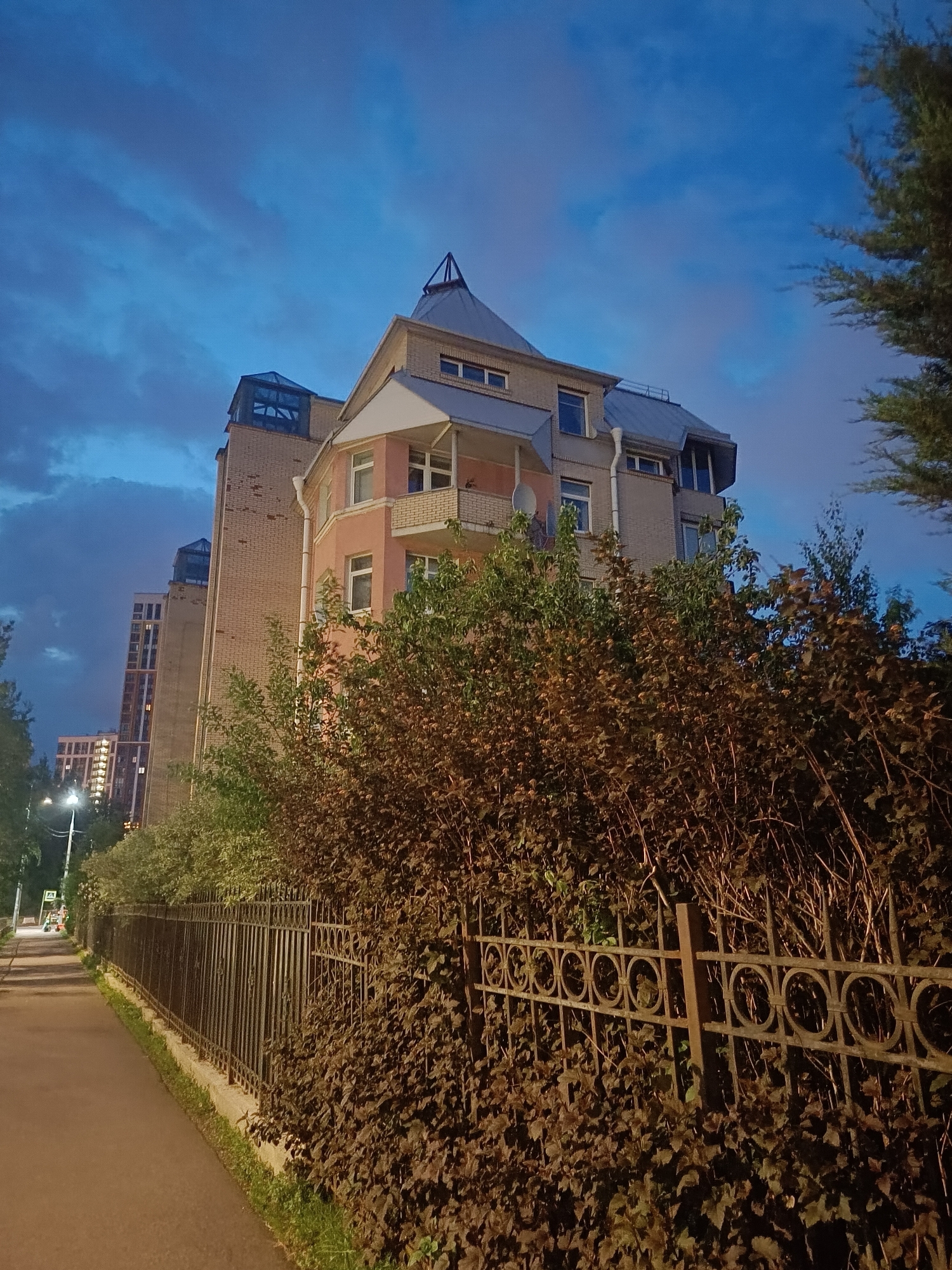 The image depicts an exterior view of a modern building in Saint Petersburg, Russia, at dusk. The building, with its distinctive architectural features including a pointed roof and a glass-enclosed upper section, is constructed primarily of brick with pink and grey accents. Adjacent to the building, a dense, overgrown bush line the front of the property. A decorative metal fence runs along the sidewalk, separating the vegetation from a paved walkway. In the background, other multi-story residential buildings are visible, their windows illuminated, suggesting an urban residential area. The sky is a deep blue, with scattered clouds, indicative of twilight. The overall scene suggests a quiet evening in a city environment.