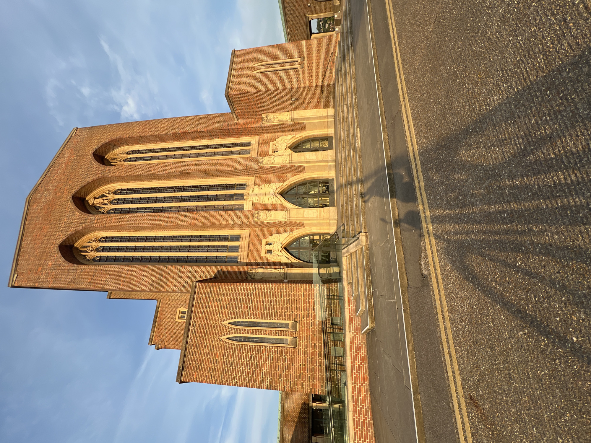 This is a vertical, eye-level, outdoor, daytime shot of Guildford Cathedral in Guildford, United Kingdom. The cathedral is made of red brick and features tall, arched windows with intricate stonework. The building is illuminated by the warm light of what appears to be a late afternoon or early morning sun, casting long shadows across the adjacent asphalt road. The road has double yellow lines and a white line marking the edge. In the background, the sky is a clear, pale blue with a few wispy clouds. The overall mood is serene and majestic, highlighting the architectural grandeur of the cathedral. No people or specific events are visible, suggesting a moment of quiet contemplation.