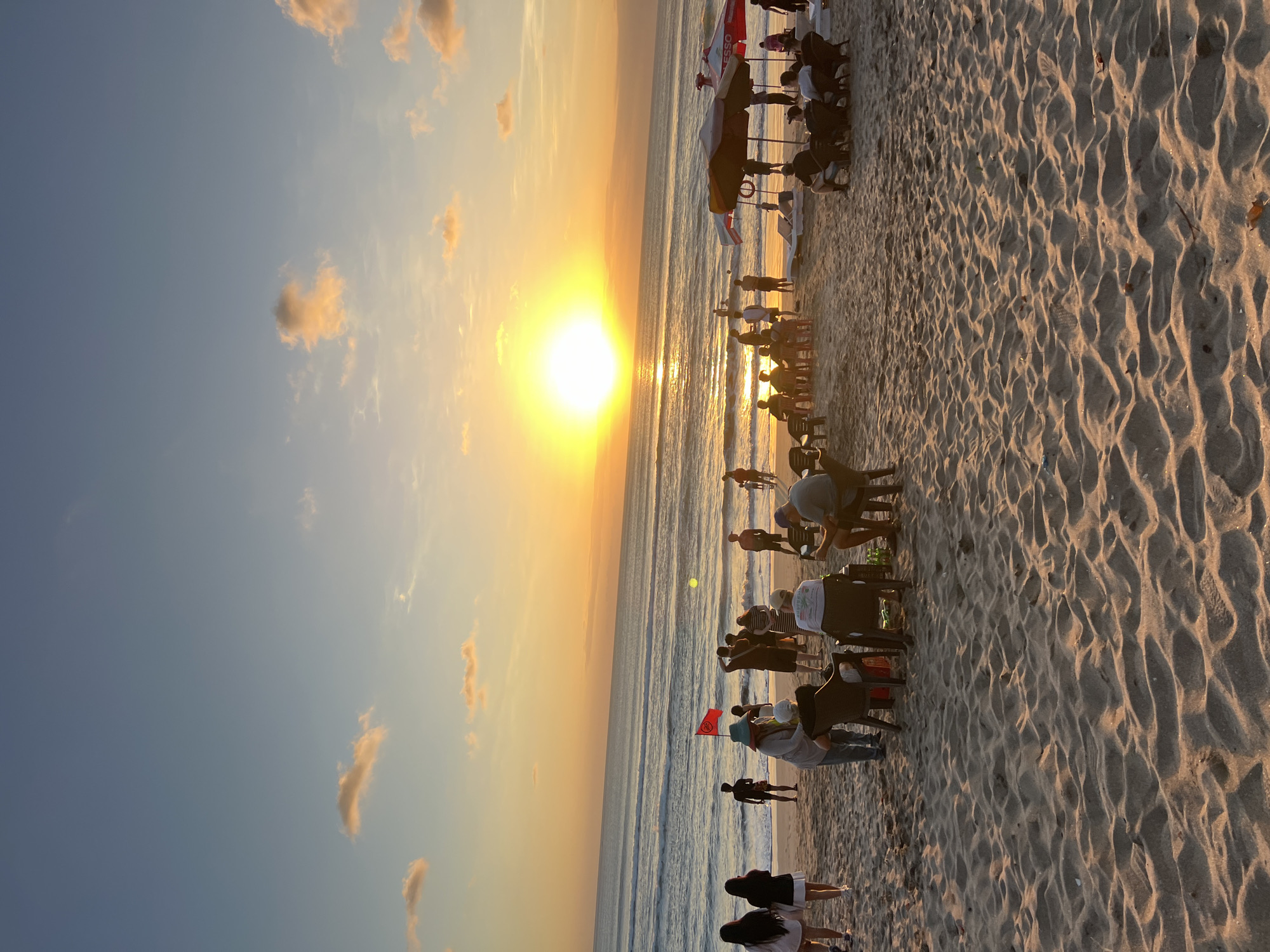 This image captures a vibrant sunset scene on Kuta Beach, Indonesia. The sky is a gradient of soft blues and oranges, with scattered clouds illuminated by the setting sun, which is positioned low on the horizon, casting a warm glow across the ocean and sand. The ocean features gentle waves rolling towards the shore, and several figures can be seen in the water.
On the beach, a diverse group of people are gathered, enjoying the twilight. Some individuals are standing, while others are seated in chairs or on loungers. A red flag, possibly a warning or marker, is planted in the sand near the water's edge. Two beach umbrellas are set up, providing shade. In the foreground, the sand is textured with ripples and shadows from the low-angled sunlight. The overall atmosphere is one of relaxation and enjoyment of the natural beauty of a Balinese sunset.