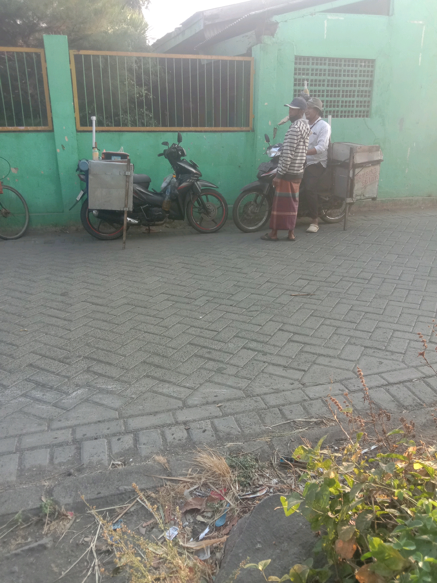 Two men are standing next to two motorcycles parked on a herringbone brick paved area. The motorcycles are black and equipped with cargo boxes. One motorcycle has a white cylindrical object attached to its cargo box. The men appear to be conversing. They are positioned in front of a green painted building with a yellow striped fence. The building has a window with a grid pattern. The scene is illuminated by sunlight, casting a shadow. The location is the city of Selotambak, Indonesia.