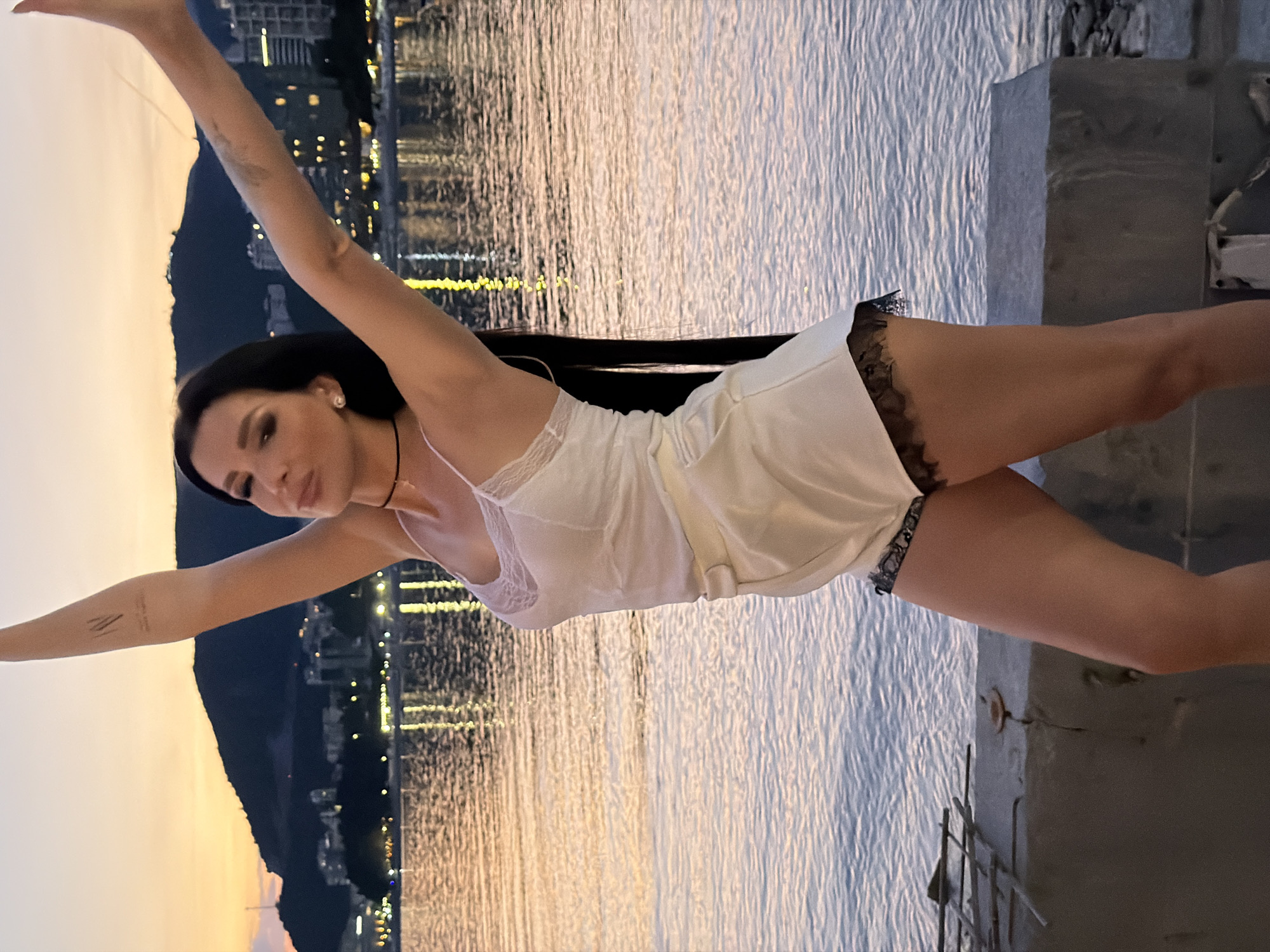 A woman is posing by the sea at sunset in Rafailovići, Montenegro. She is wearing a white nightgown with lace trim and has long black hair. Her arms are outstretched. The backdrop shows a coastline with buildings lit up at dusk, reflecting in the water. The setting appears to be a harbor or waterfront area with concrete structures visible on the right side of the frame.