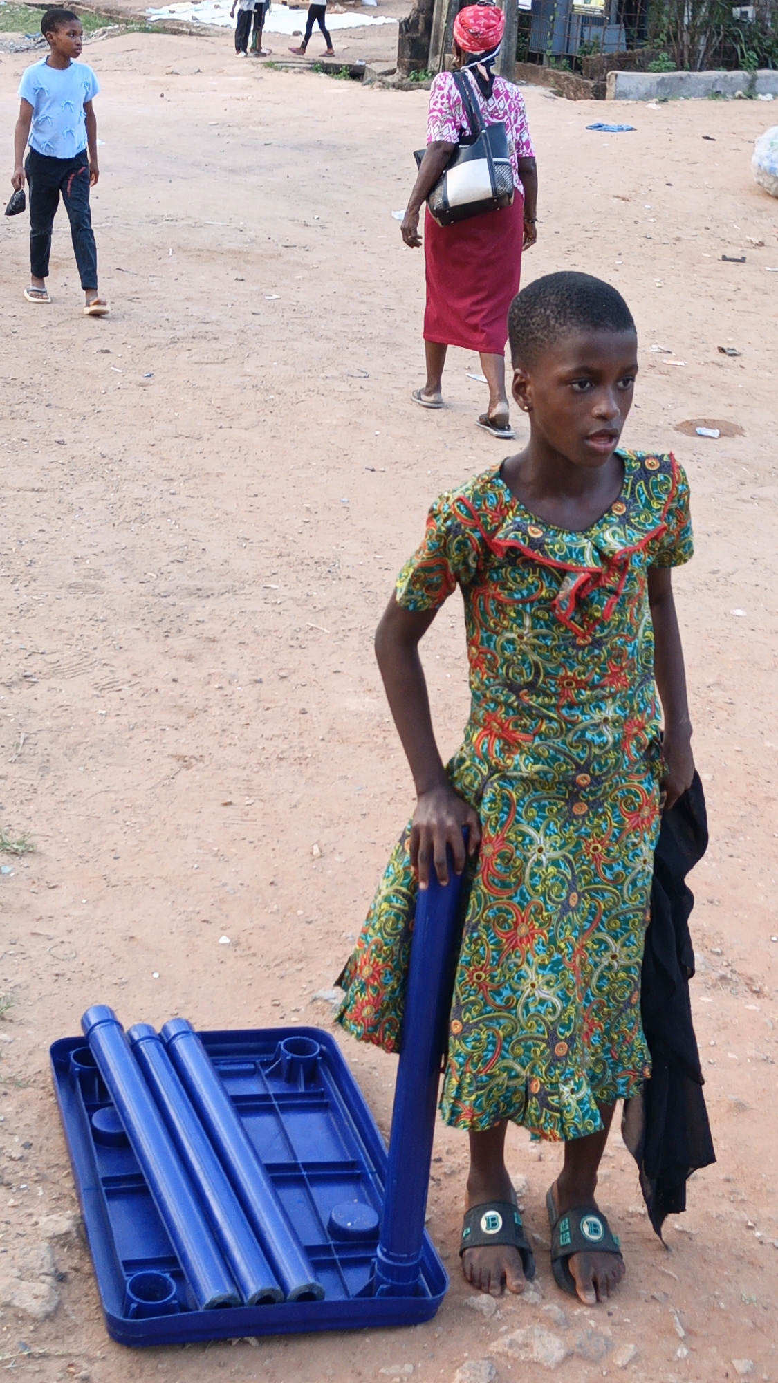This outdoor image captures a scene on a reddish-brown, unpaved dirt path in what is identified as Onitsha, Nigeria. In the foreground, a young Black girl stands, gazing slightly to her right with her mouth partially open. She is dressed in a vibrant, short-sleeved dress featuring a busy pattern of green, red, and blue swirls and floral designs. Her left hand rests on a blue plastic cylindrical pole, while a blue plastic tray lies beside her on the ground, containing three similar blue poles. These objects appear to be components of a disassembled small table or stool. She also holds a piece of black fabric in her left hand. On her feet, she wears black slide sandals, each strap adorned with a white logo, possibly "LD", set within a small green rectangle.
In the mid-ground, other people are seen walking along the same path. To the left, a young Black boy in a light blue t-shirt and dark pants walks away from the camera, carrying a small black object. On the right, a Black woman, viewed from behind, is walking away. She wears a red headscarf, a patterned top with pink and white, and a long red skirt, carrying a black and white shoulder bag. Additional indistinct figures are visible further down the path, and rudimentary structures with some vegetation can be seen in the background. The lighting suggests it is daytime with natural, soft illumination. No explicit text is visible beyond the logos on the girl's sandals. The scene depicts everyday activity in a simple, unpaved setting.