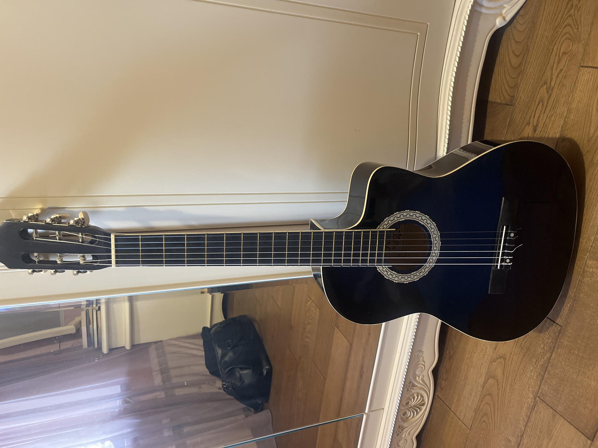 An acoustic guitar stands upright on a wooden floor. The guitar is dark blue and black, with six strings and a soundhole surrounded by a decorative rosette. It leans against a plain, light-colored wall. To the left of the guitar is a large, ornate white mirror with a detailed frame. Reflected in the mirror are sheer curtains, a radiator, a dresser with books stacked on top, and a black duffel bag lying on the floor. The scene appears to be indoors, possibly in a bedroom or living area. There are no people visible in the image. The lighting suggests it is daytime, with light coming from a window behind the curtains, as seen in the reflection.