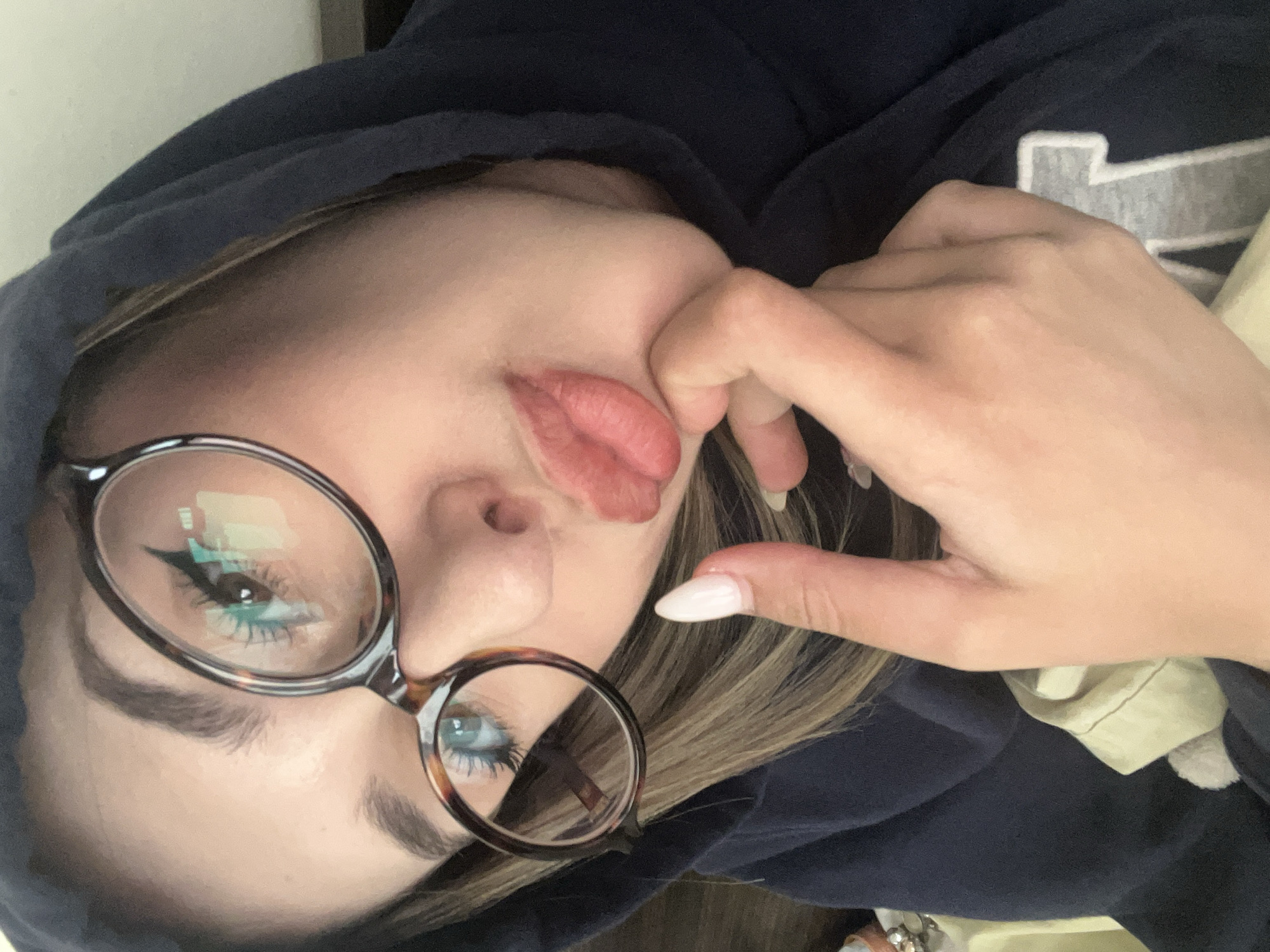 A woman with fair skin and light brown hair wearing glasses and a dark blue hooded sweatshirt is pictured in a selfie-style shot. Her lips are slightly pursed. She has a white-tipped, oval-shaped fingernail visible, and her hand is positioned near her face. The reflections in her glasses reveal a light-colored interior with what appears to be furniture. The location cannot be determined from the image.