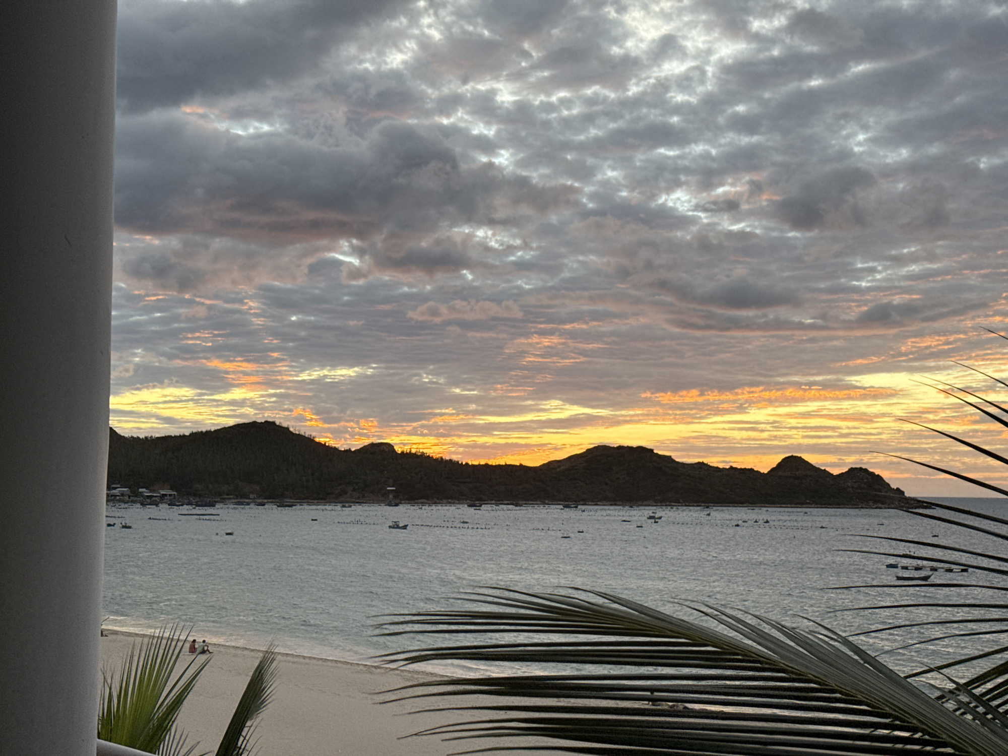 A beachfront scene at sunset is captured from a balcony, with a white column on the left partially obscuring the view. The sky is filled with dramatic, textured clouds, tinged with orange and yellow from the setting sun. Below the sky is a dark, undulating mountain range bordering the coast. The water is a calm, muted blue-green, with numerous small boats and what appear to be buoys or floating structures scattered across it, indicating a harbor or aquaculture farm. The foreground features a sandy beach, with two figures sitting near the water's edge, and the fronds of a palm tree in the bottom right and left corners of the frame, adding a tropical feel. The overall mood is serene and picturesque.