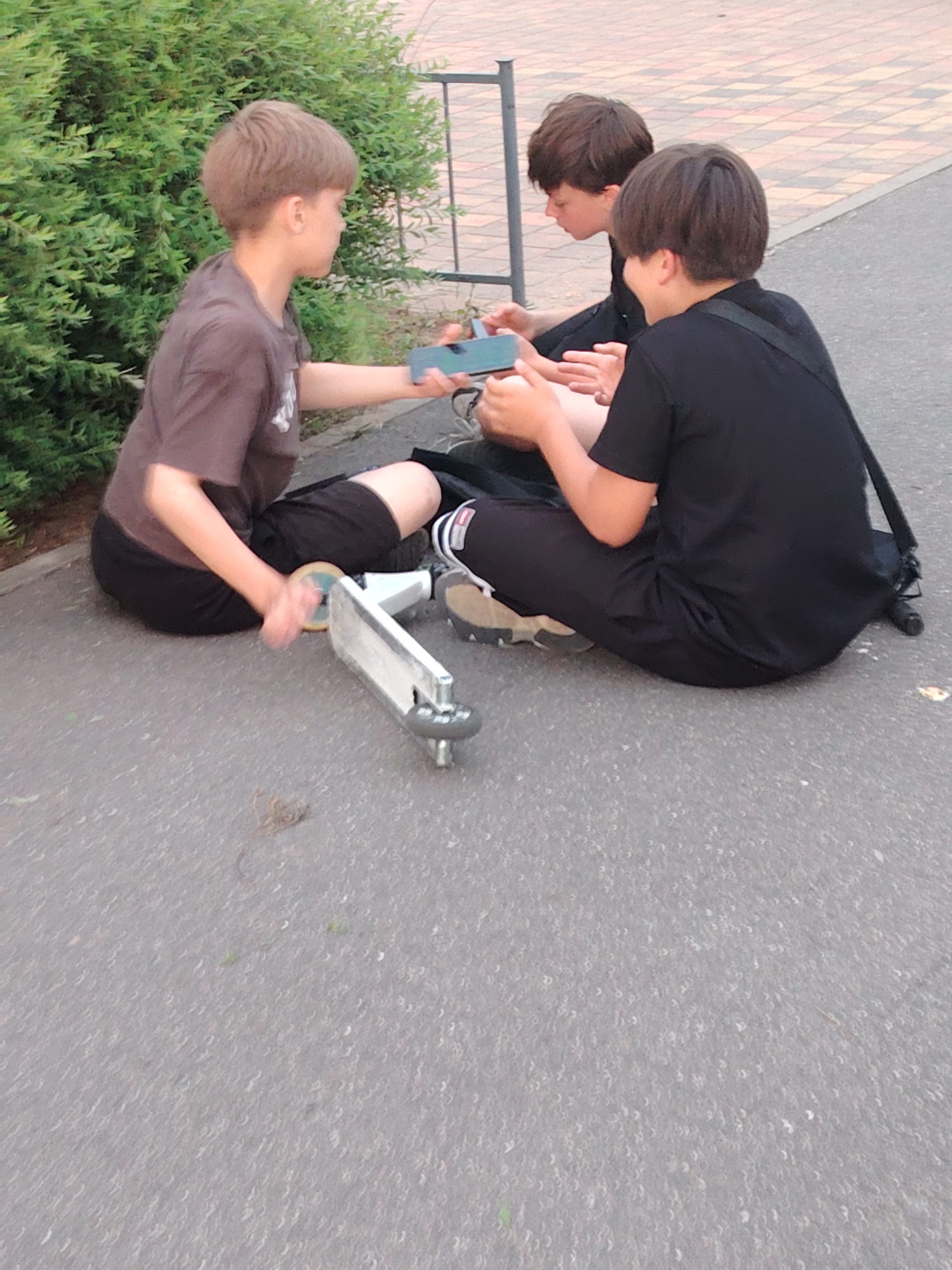 Three young males are seated on a grey paved surface in Бараўлянскі сельскі Савет, Belarus. The male on the left, with light brown hair, wears a dark brown t-shirt and dark shorts, holding a blue smartphone in his right hand, extended towards the male in the center. A white scooter lies horizontally on its side on the ground near his left. The central male, with dark brown hair, wears a dark t-shirt and dark shorts, looking down at the presented smartphone. The male on the right, with darker brown hair, wears a black t-shirt and black shorts, with a black strap over his right shoulder, observing the interaction. The ground is a textured grey asphalt or similar paving, with visible small debris. To the immediate left is a dense green bush. In the mid-ground, a metal fence separates the paved area from a tiled path with a light and dark checkered pattern. The individuals appear engaged in an activity involving the smartphones.