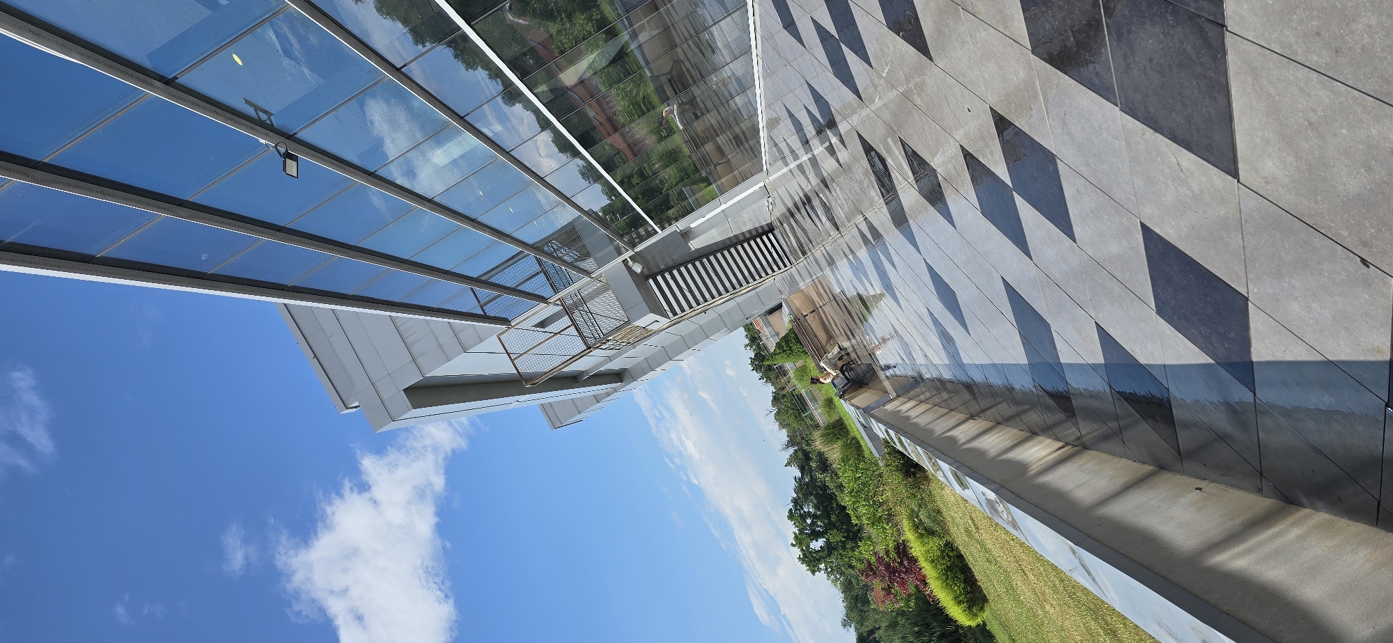 The image displays a modern architectural scene captured from a low angle, looking upwards towards a building within the International Fair Plovdiv complex in Plovdiv, Bulgaria, under a clear blue sky with scattered white cumulus clouds.
The building on the right features a multi-story glass curtain wall, reflecting the vivid blue sky, white clouds, green foliage, and what appears to be the roofline of a distant, older, red-tiled building. Water droplets and streaks are visible on the glass surface. A black, rectangular outdoor light fixture is affixed to one of the glass panels. The left side of the building exhibits a sleek, light grey paneled facade. An external metal staircase with a mesh railing, finished in a bronze or gold tone, descends from the building's mid-section on this side.
The foreground and mid-ground are covered by a highly reflective paved area, composed of large, diamond-shaped tiles arranged in an alternating pattern of light grey and dark grey/black. This surface is wet, showing strong reflections of the sky and light, emphasizing the checkerboard pattern. In the mid-ground, on the tiled patio, a person wearing light-colored clothing is seated on a low bench or wall, facing away from the camera, looking towards the landscaped area. A light-colored dog is standing or sitting directly next to the person.
To the far left of the paved area, a vibrant green landscape unfolds, featuring well-maintained grass, various shrubs, and trees, including some with reddish foliage, providing a natural backdrop to the contemporary architecture. The scene is brightly illuminated by natural sunlight, creating strong reflections and subtle shadows.