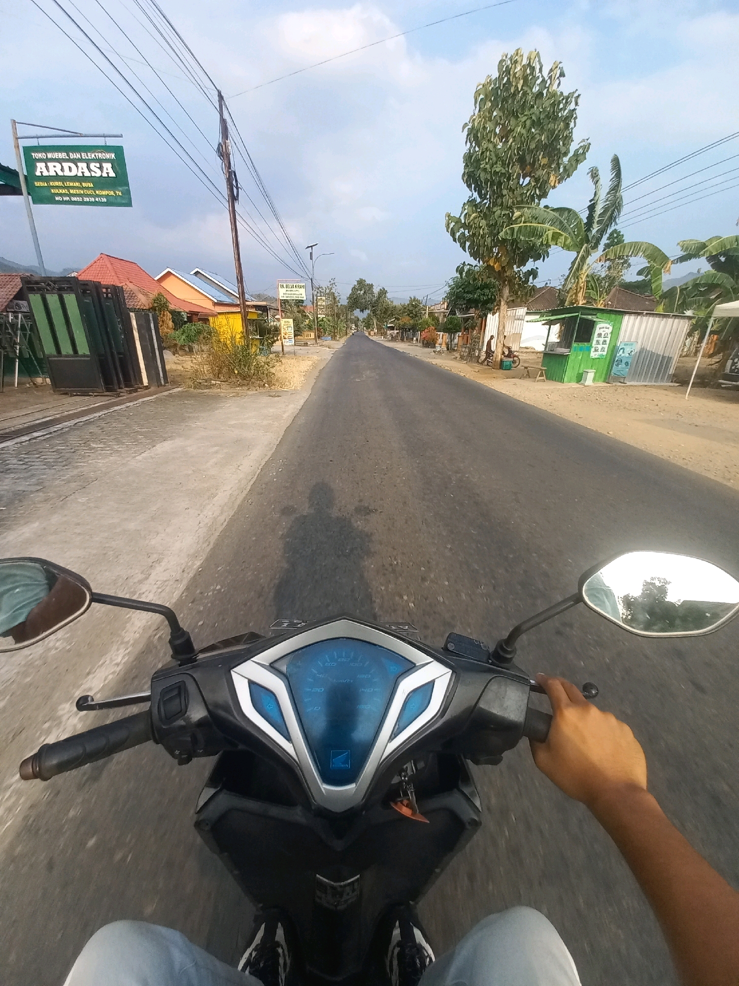 The image captures a first-person perspective of someone riding a motorcycle on a paved road in Ngarjosari, Indonesia. The motorcycle's handlebars, dashboard, and a portion of the rider's hand and legs are visible in the foreground. The road extends into the distance, lined with small buildings, trees, and utility poles. A green sign for "ARDASA" which appears to be a furniture and electronics store, is prominently displayed on the left side of the road. The sky is partly cloudy. The overall impression is that of a typical rural or suburban scene in Indonesia.