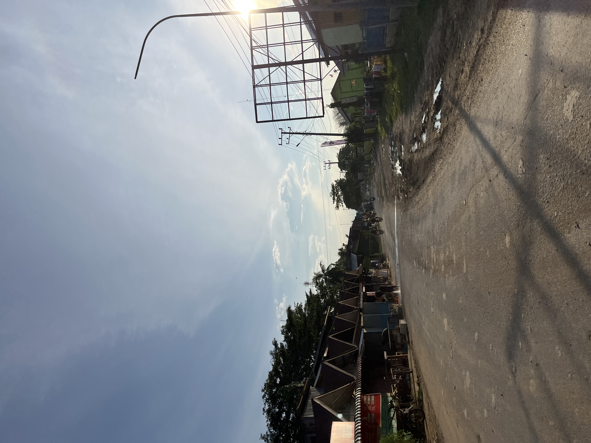 The image depicts a dusty, unpaved road in Alurcucur, Indonesia, lined with small, modest buildings and shops. The sky is a bright blue with scattered clouds and the sun is shining, casting long shadows. Several motorcycles are parked along the side of the road, and a few are visible in motion. Some of the shops appear to be selling goods, with items displayed outside. A person is seen hosing down the road in front of one of the shops. There are also utility poles with electrical wires crisscrossing overhead, and a large, empty billboard structure. Puddles of water and mud are present on the road surface, indicating recent rain or poor drainage. The overall impression is of a busy, yet somewhat dilapidated, rural or semi-urban street scene.