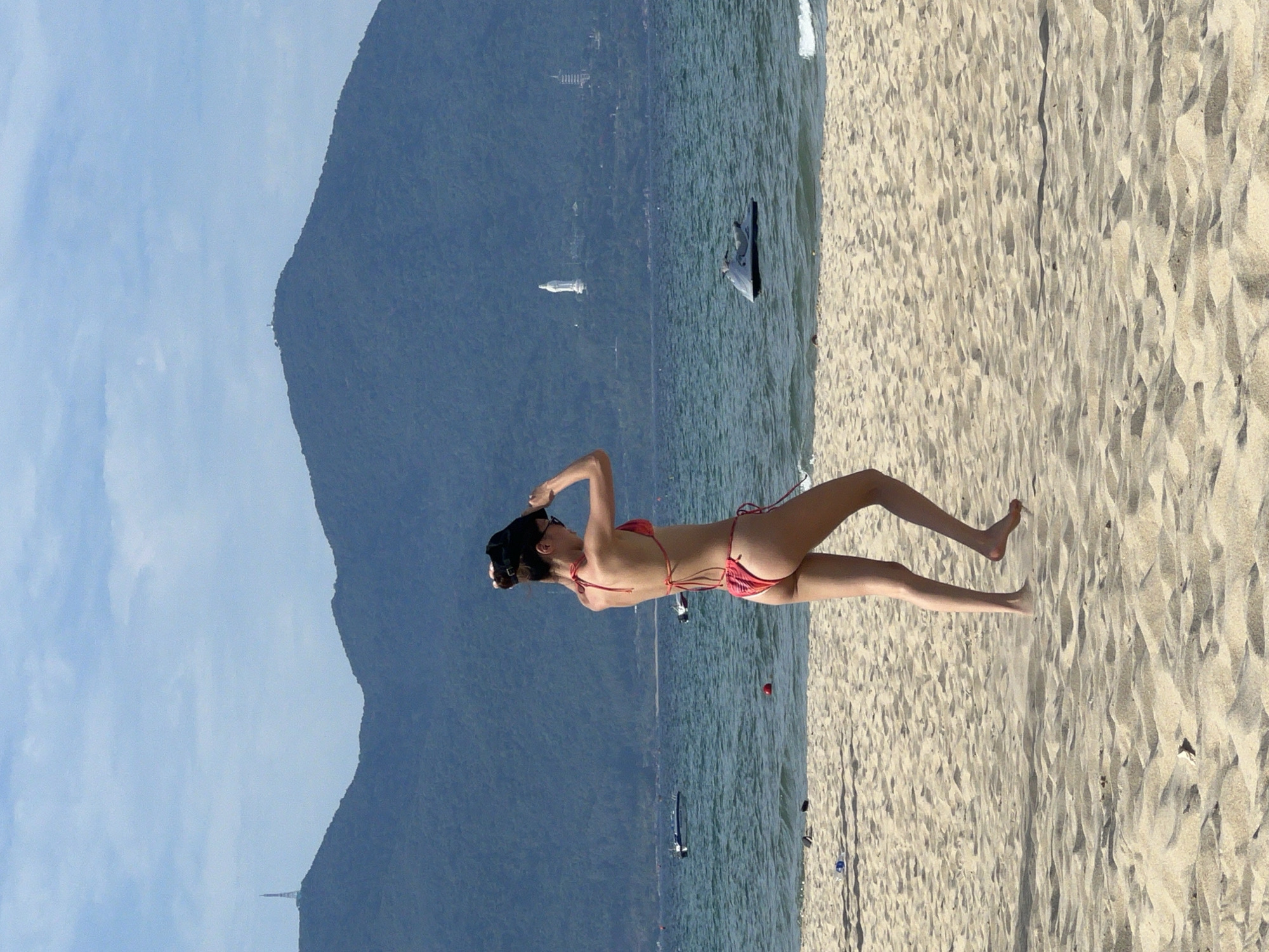 A woman in a red bikini is walking on a sandy beach in Da Nang, Vietnam. She is holding a black object, possibly a hat, above her head. In the background, the sea is visible with a few boats and a jet ski. Beyond the sea, there are lush green mountains with some structures on them, including a white statue of a woman and a tall, slender tower. The sky is partly cloudy. The scene depicts a sunny day at the beach with a view of the coastal landscape of Da Nang.