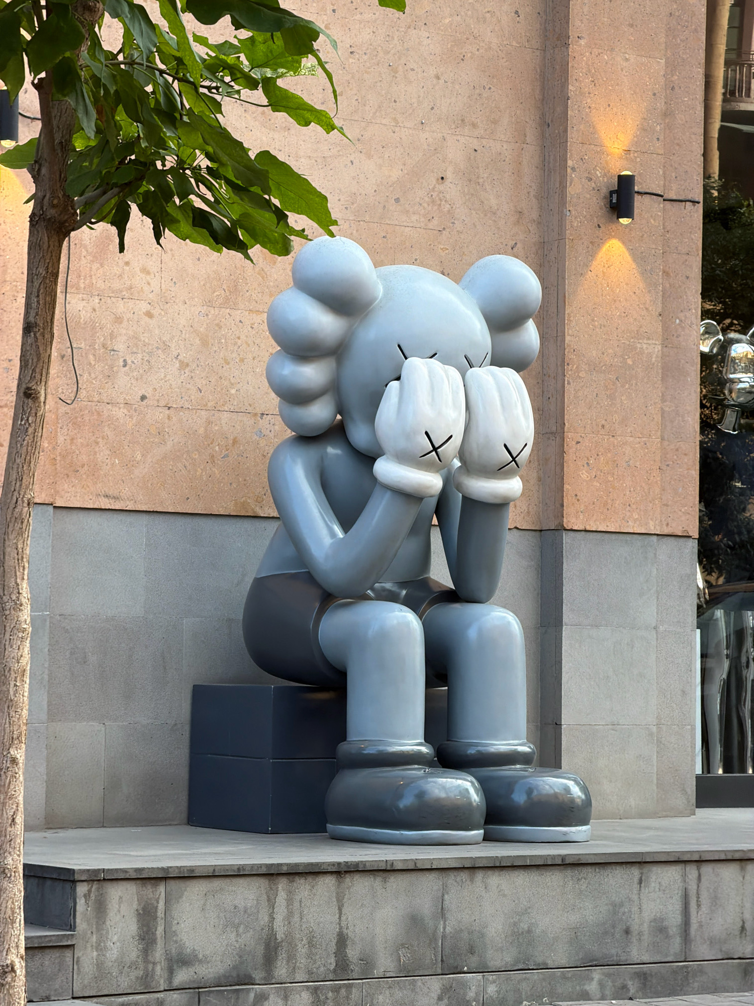 A grey, cartoonish sculpture of a figure with its hands over its eyes sits on a dark grey cube in front of a building with a tan, stone-tiled facade. The sculpture is in a seated position, with its knees bent and its feet flat on the ground. The building has a black outdoor light fixture with two illuminated bulbs mounted on its facade. To the left of the sculpture, a tree trunk and green leaves are visible. The ground in front of the sculpture is paved with grey stone tiles. To the right of the sculpture, a reflection of a silver, cartoonish figure is visible in a shop window. The scene appears to be an outdoor public space in Yerevan, Armenia.