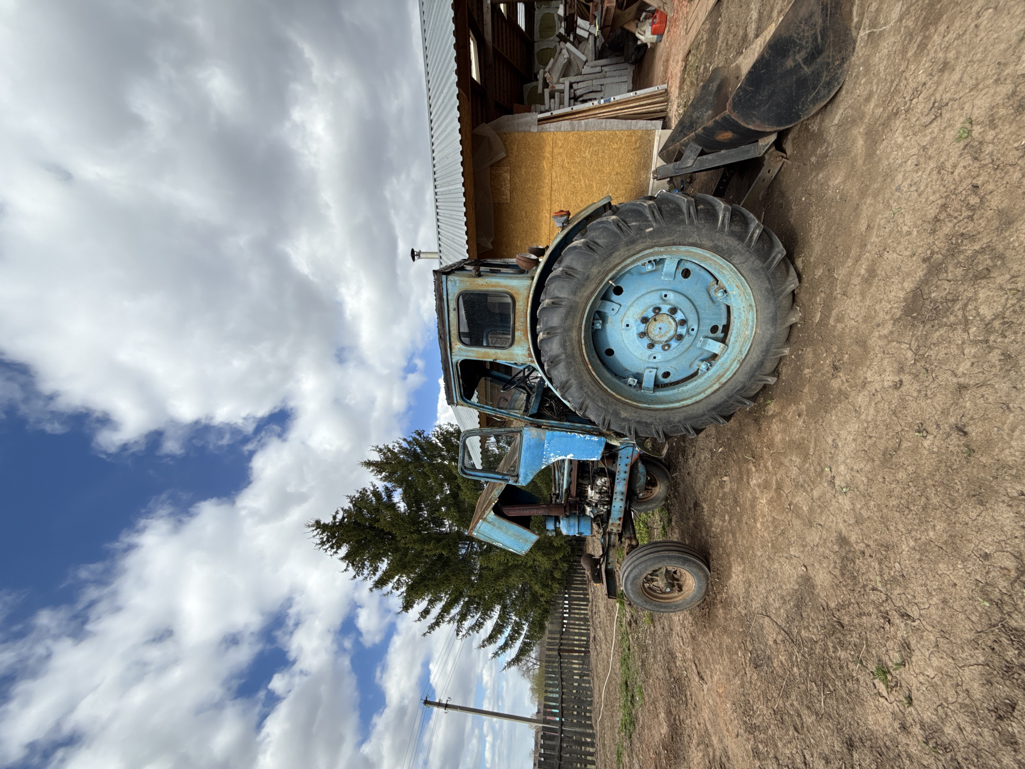 The image shows a blue, dilapidated tractor situated outdoors. The tractor appears to be in disrepair, with rust and missing components. Adjacent to the tractor is a wooden shed or structure with a corrugated metal roof. Scattered debris and equipment are visible within the shed. In the background, a partially obscured pine tree stands near a wooden fence and utility pole. The sky is cloudy, and the ground is dirt and grass. The image was taken in Nizhnyaya Chermoda, Russia.