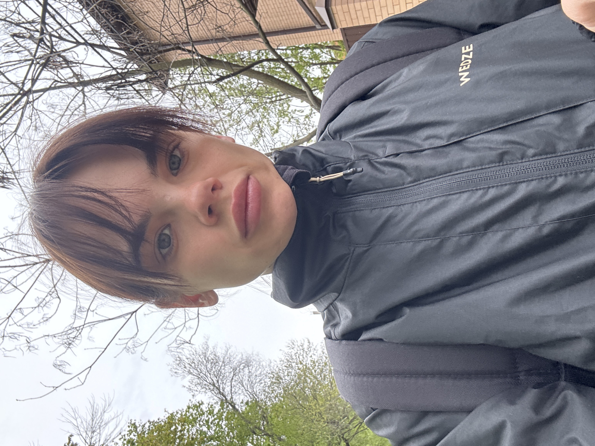 The image is a close-up, low-angle selfie of a young person captured outdoors. The subject, positioned centrally and rotated ninety degrees clockwise, has short, dark hair with bangs, light-colored eyes, and a neutral expression while looking directly at the camera. They are wearing a dark, possibly black or navy, hooded jacket with a visible zipper down the front. The brand name "WEDZE" is printed in light letters on the left side of the jacket. A strap from a dark backpack is visible on their right shoulder.
The background features an overcast sky, indicating a cloudy daytime. Several trees are visible; some have bare branches, while others show early signs of green leaves, suggesting the season is spring. A structure made of light brown brick is partially discernible behind the trees in the upper left portion of the frame. The scene conveys an outdoor setting, potentially a park or urban green space in Rostov-on-Don, Russia. The overall lighting is soft and even.