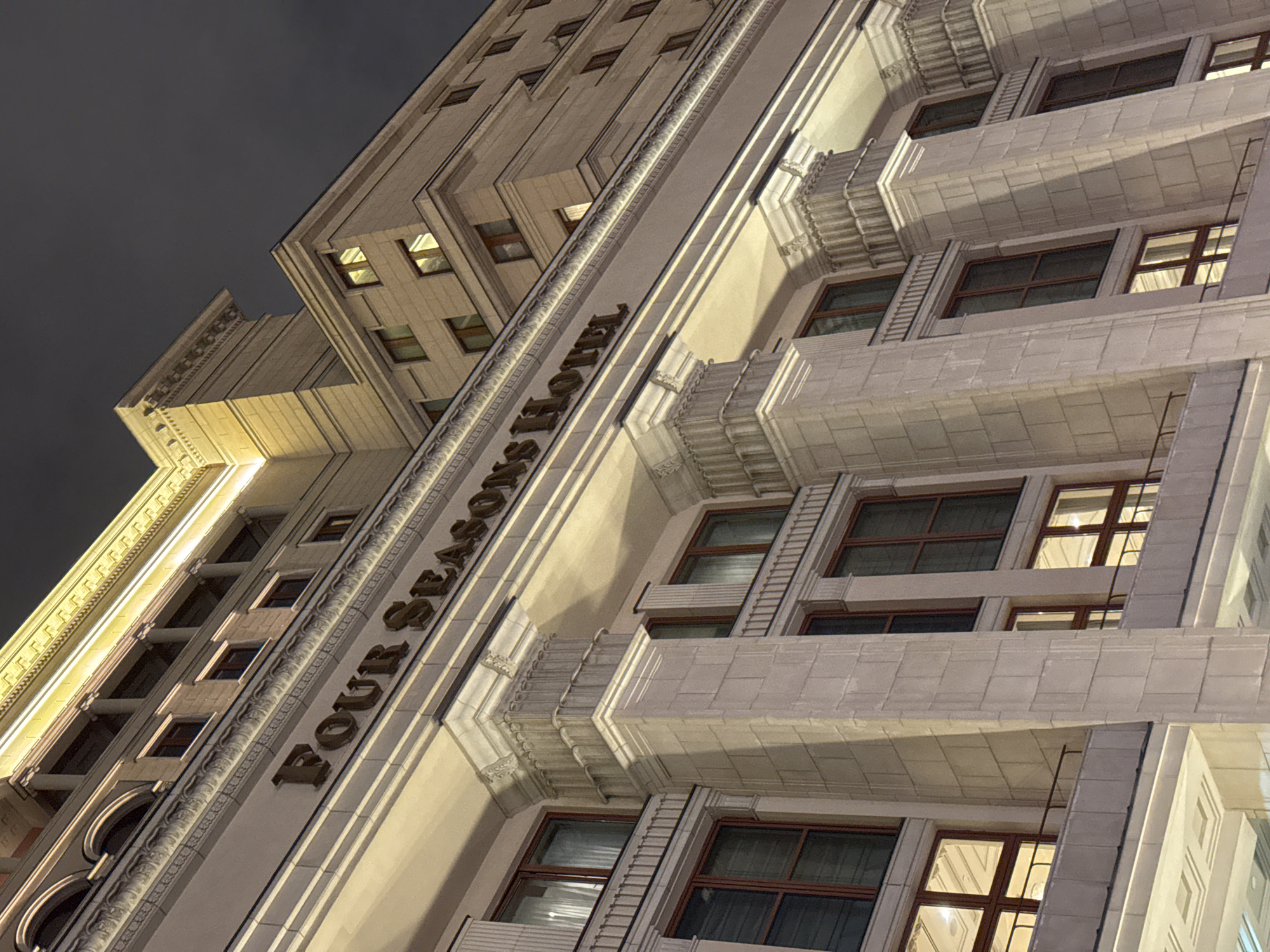 This is a low-angle nighttime shot of the Four Seasons Hotel in Moscow, Russia. The building is an architectural composition of light stone featuring a series of stacked, stepped-back sections. The hotel's name is displayed vertically in prominent letters along one of the building's facets. The windows are dark, though a few show interior light, indicating occupancy. The sky is overcast. The architectural style is monumental, with heavy emphasis on vertical lines and classical ornamentation.