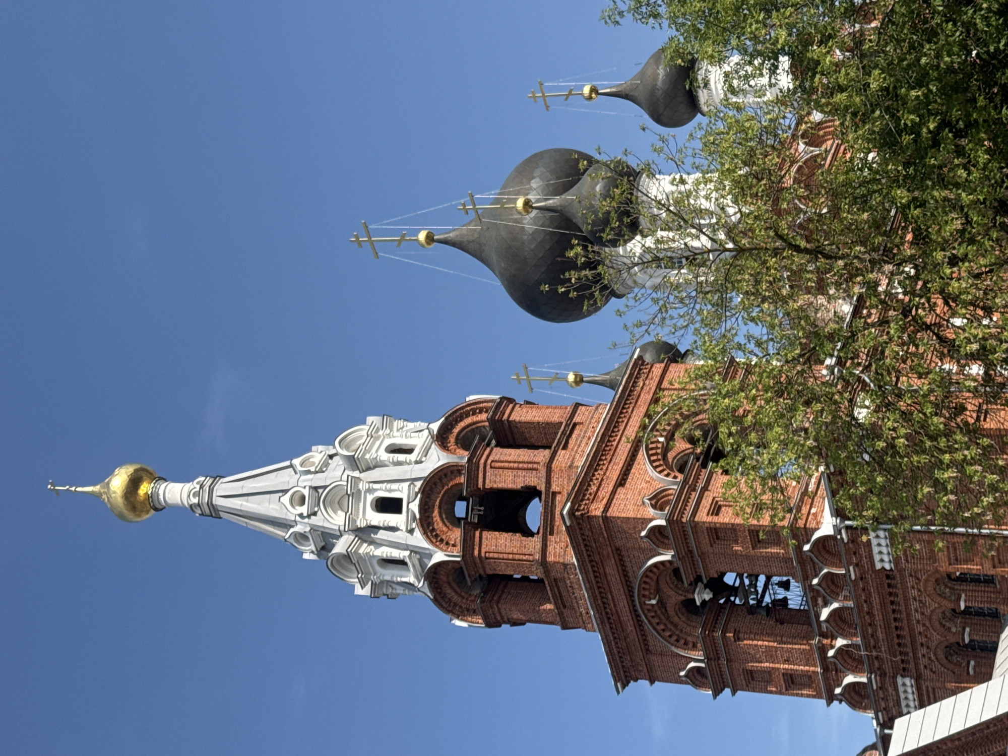 The image shows a close-up view of the bell tower and domes of a Russian Orthodox church against a clear blue sky. The bell tower is constructed of red brick with white decorative elements and arched openings. A golden dome topped with a cross is visible above the bell tower. To the right of the bell tower are several black onion domes, also topped with crosses. Green foliage from trees obscures parts of the lower sections of the church. The architecture suggests a traditional Russian church design. The location is identified as Nizhny Novgorod, Russia.