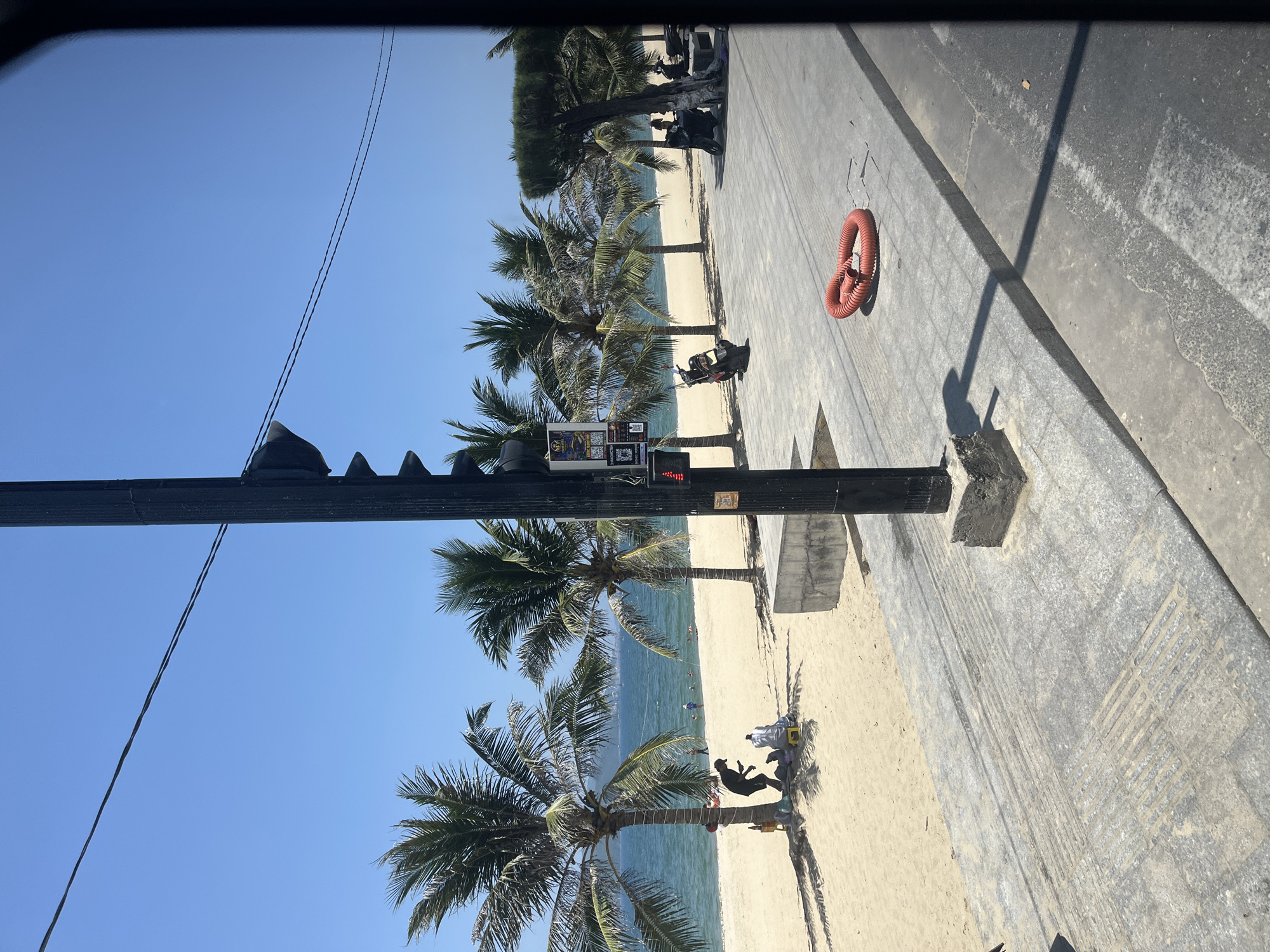 The image depicts a beach scene in Nha Trang, Vietnam, on a clear, sunny day. In the foreground, a paved walkway runs parallel to the beach, with a black lamppost situated on the right side of the frame. A bright orange, coiled hose lies on the pavement. Beyond the walkway is a sandy beach lined with palm trees. Several people are visible on the beach, some seated under the shade of the palm trees, and others in the shallow water or further out in the ocean. The sea is a vibrant blue, meeting the horizon under a clear sky. Two motorcycles are parked on the paved area to the right. A traffic light signal is attached to the lamppost.
