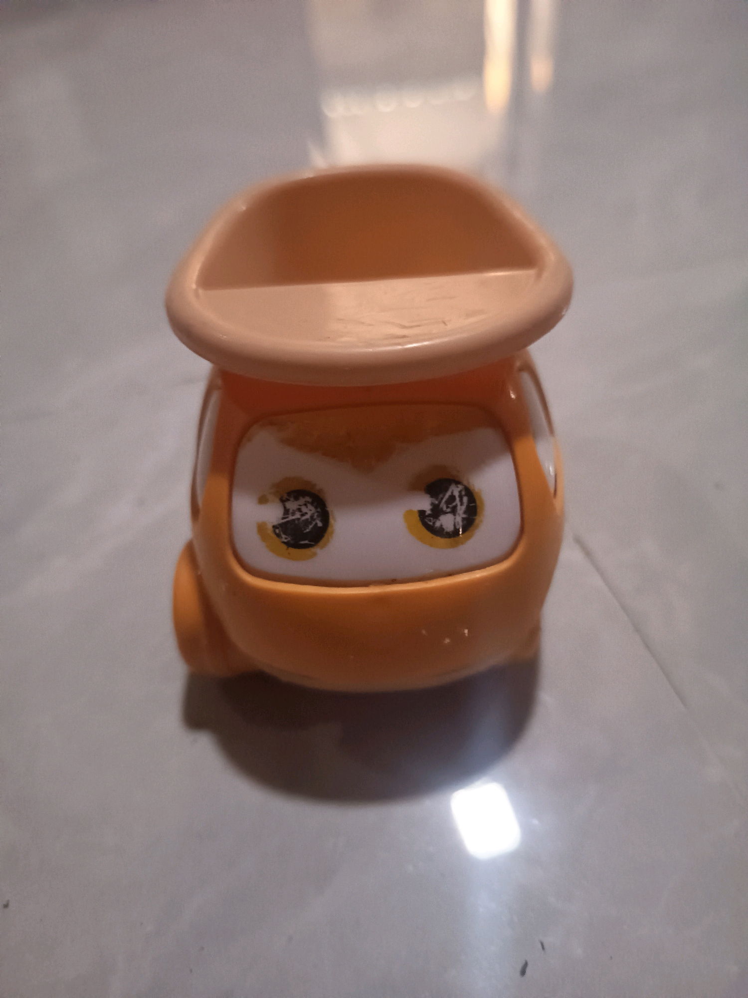 A close-up, eye-level, front-facing shot captures an orange plastic toy on a light gray, tiled surface. The toy resembles a stylized car with a wide, light orange, bowl-shaped top. The front of the toy features two large white oval "eyes" with black pupils outlined in yellow. The pupils are smudged, with some black material flaking off. The toy has a rounded, bulbous body and appears to be sitting on a flat surface. The lighting creates a bright, rectangular reflection on the surface beneath the toy. No people or specific city locations are visible.