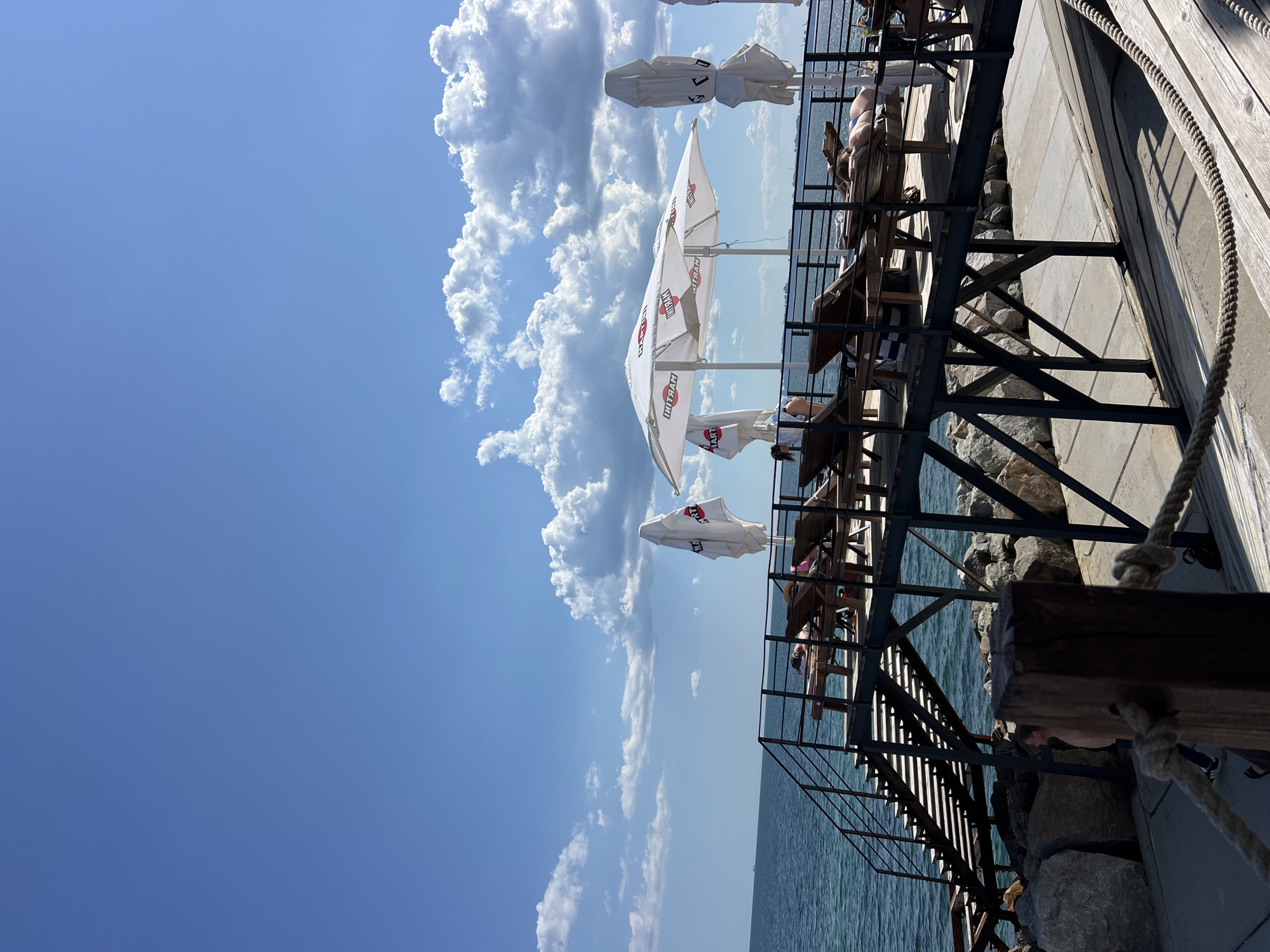 The image depicts a pier on the Black Sea coast in Fontanka, Ukraine, on a sunny day with scattered clouds. The pier is constructed of wood and metal, featuring a staircase leading up to a platform with several empty and occupied lounge chairs. Two white umbrellas, one open and displaying the "MARTINI" logo, are set up on the pier. Several people are visible on the lounge chairs, some appearing to be sunbathing. The sea is calm and blue, with a faint outline of ships visible on the horizon. The pier is supported by a metal structure and a foundation of large rocks. A wooden post with a rope is in the foreground, partially obscuring a concrete walkway.