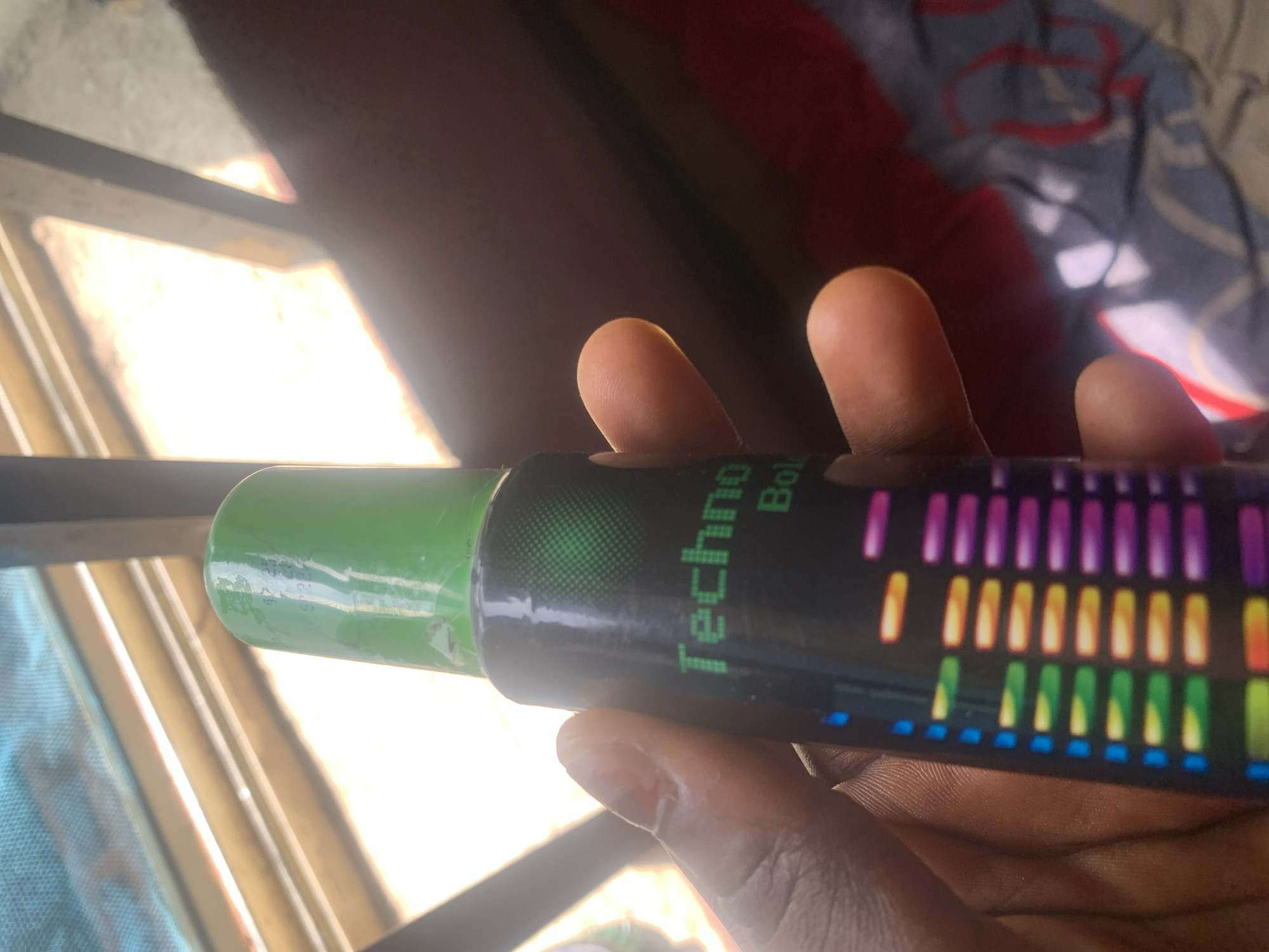 A person's hand holds a black aerosol can with a green cap. The can features colorful bars resembling an equalizer display and text that reads "Techno Bol." The background shows a window with natural light filtering through and a blurred red and grey patterned fabric. The scene is likely indoors, near a window.