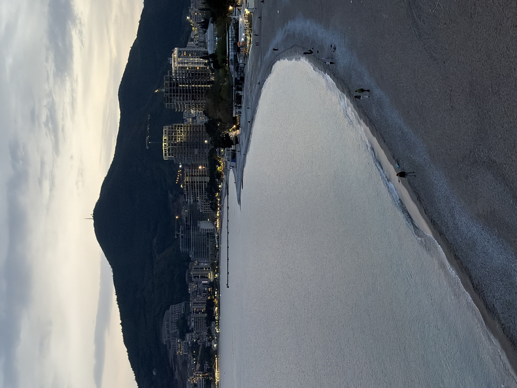 The image depicts a nighttime view of a beach in Budva, Montenegro. A dark, tree-covered mountain rises along the left side of the image. Buildings with illuminated windows are situated between the mountain and the beach. The beach extends along the right side of the image with the Adriatic Sea occupying a significant portion. Several small figures of people are walking or standing on the shoreline. The sky is overcast with grey clouds.
