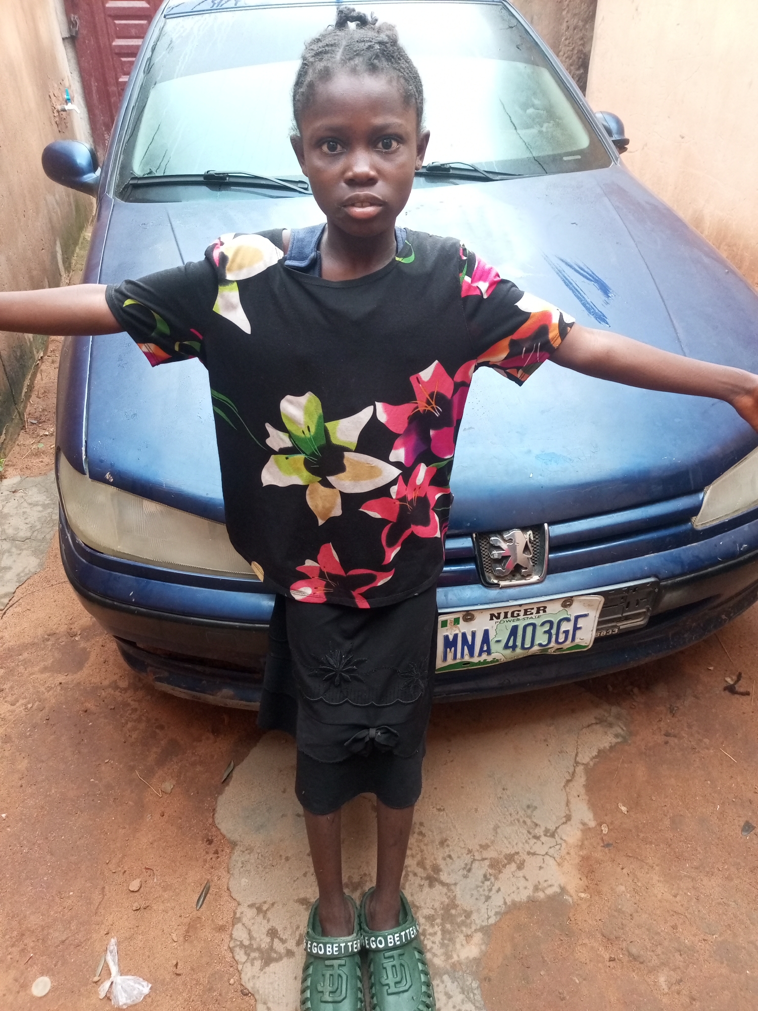 A young girl stands in front of a blue car in what appears to be a residential area in Tempure, Nigeria. The car has a license plate that reads "MNA-403GF". The girl is wearing a black t-shirt with a floral pattern and a black skirt. She is also wearing green sandals with the words "GO BETTER" written on them. The ground is a mix of dirt and concrete. The environment appears to be an outdoor setting with a building visible in the background.