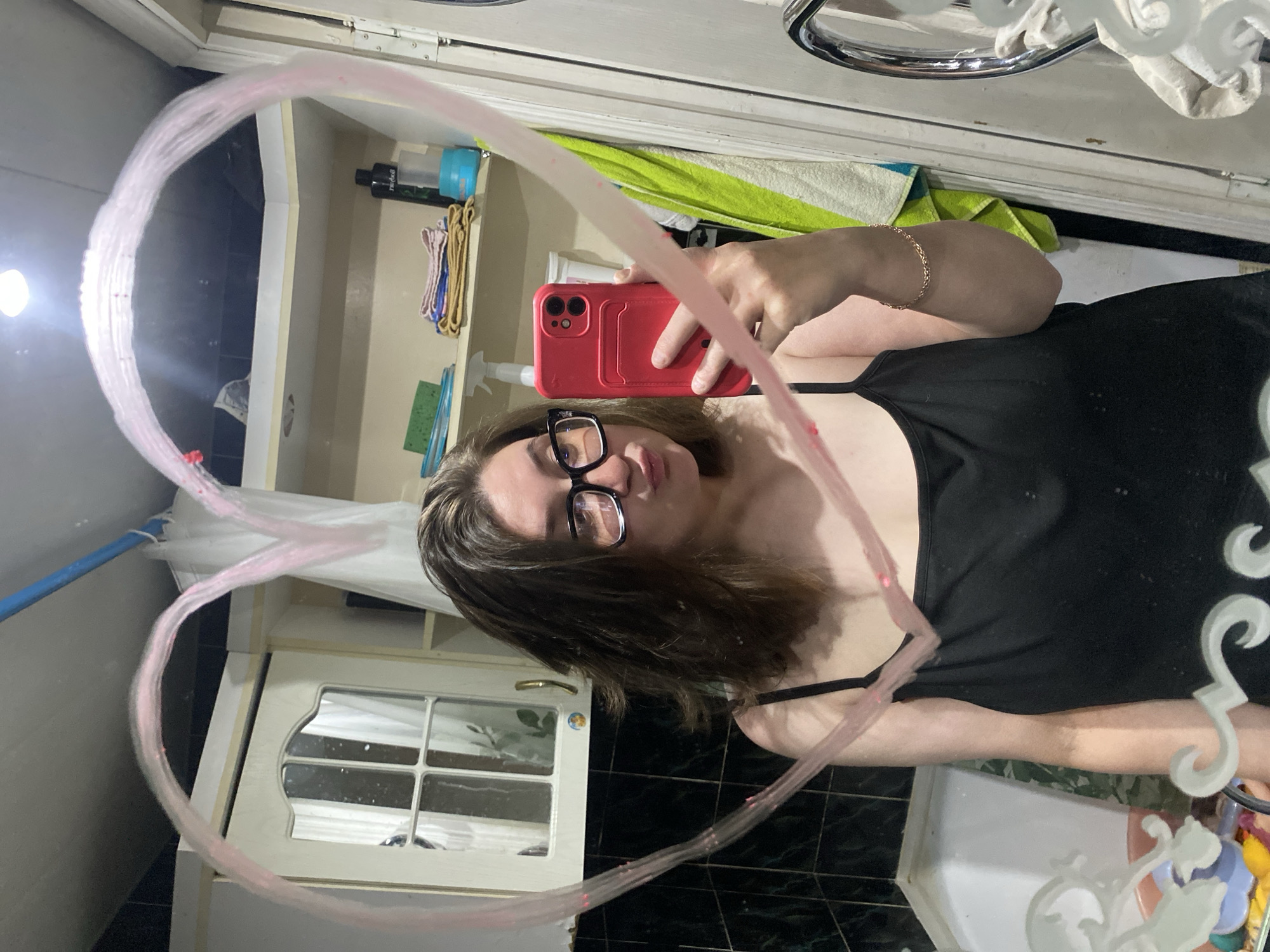 A person is taking a selfie in a bathroom mirror. The person has dark hair and wears glasses and a black tank top. They hold a red phone in their right hand. A heart is drawn on the mirror in pink. The bathroom includes white cabinets, dark tile flooring, and various toiletries. A green towel is visible on a shelf. It is not possible to verify the location is Chaikovsky, Russia.