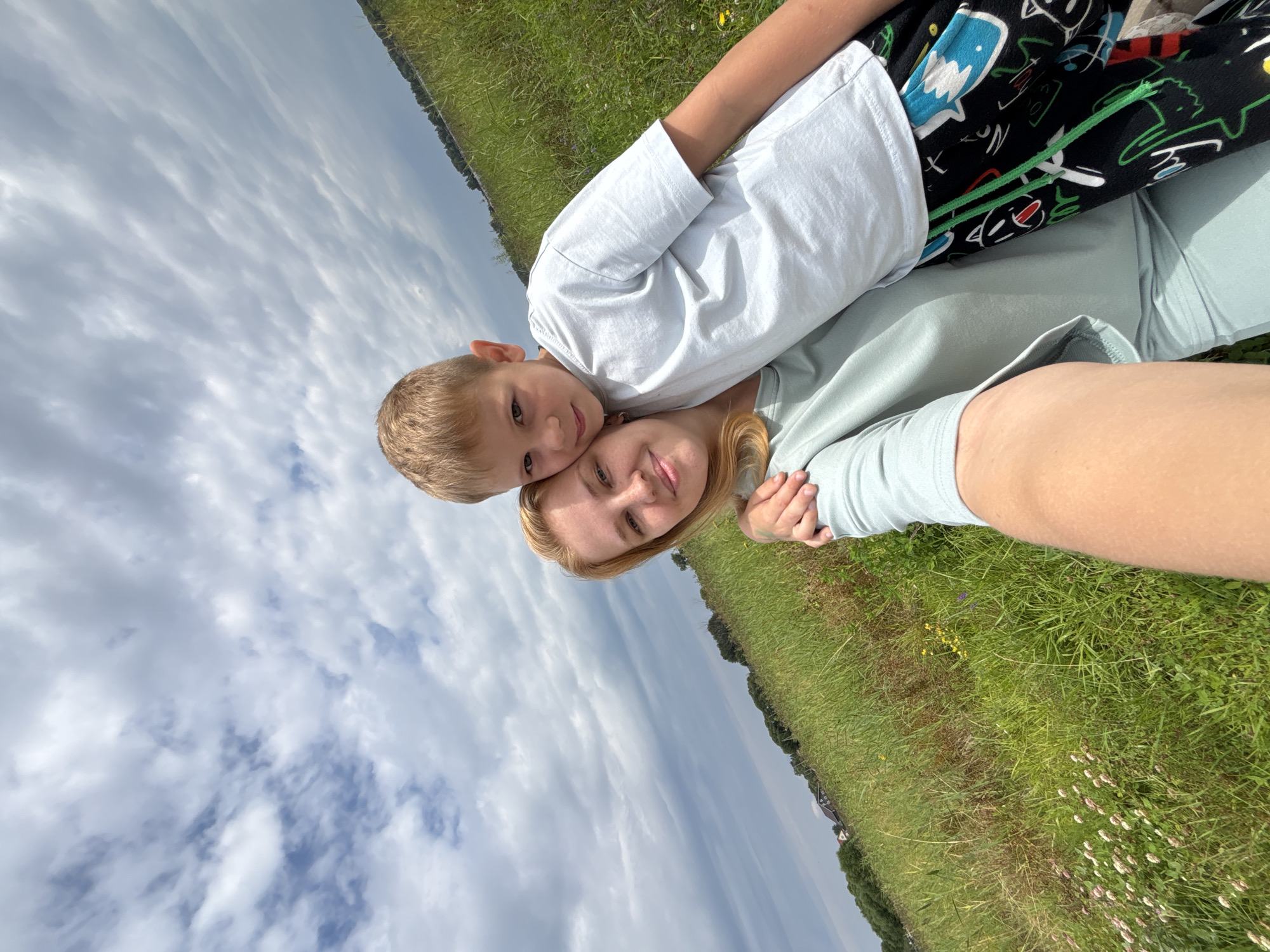 This is a selfie of a young boy and a woman lying on a grassy field. The boy, with blonde hair and blue eyes, is lying on top of the woman, with his head resting on her forehead. He is wearing a white t-shirt and patterned shorts. The woman has blonde hair and is wearing a light blue shirt. They appear to be enjoying themselves, with the woman smiling slightly and the boy looking at the camera.
The background consists of a vast expanse of green grass, with some wildflowers scattered throughout. In the distance, trees and some buildings can be seen, suggesting a rural or park-like setting. The sky is cloudy, with a mix of blue sky and white clouds, indicating it is daytime. The overall mood of the image is cheerful and relaxed, capturing a moment of connection between the two individuals.
The image appears to be taken outdoors in a natural setting, likely during the day. There is no visible text in the image.
