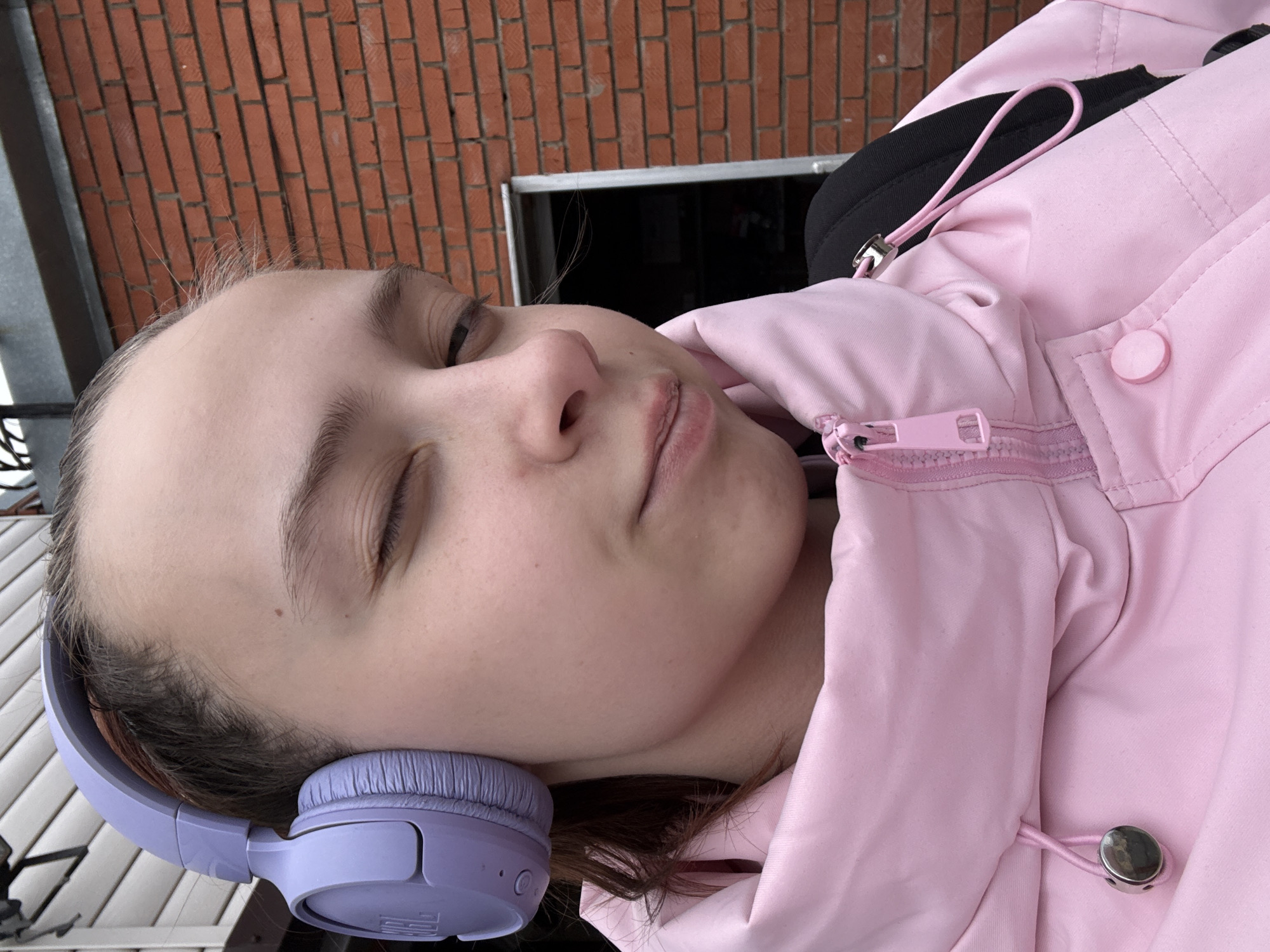 A young person is seen in a close-up, slightly off-center, portrait orientation. The individual, with their eyes closed, wears light purple headphones over their ears and is dressed in a pink jacket with the zipper partially closed. A black backpack strap is visible behind the jacket. The background features a brick wall, and a dark doorway or window is partially visible. The location is Прикамский, Russia.