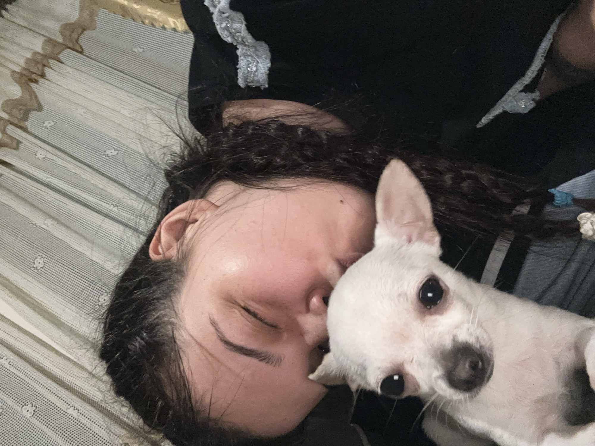 A woman with curly brown hair, lying on a patterned surface, is facing slightly to the left with her eyes closed. A small white dog with a dark nose is lying close to her, its head near her face. The dog is looking at the camera. The woman is wearing a dark-colored top with some trim. The location is not evident in the image to determine it is in Chubra, Bulgaria.