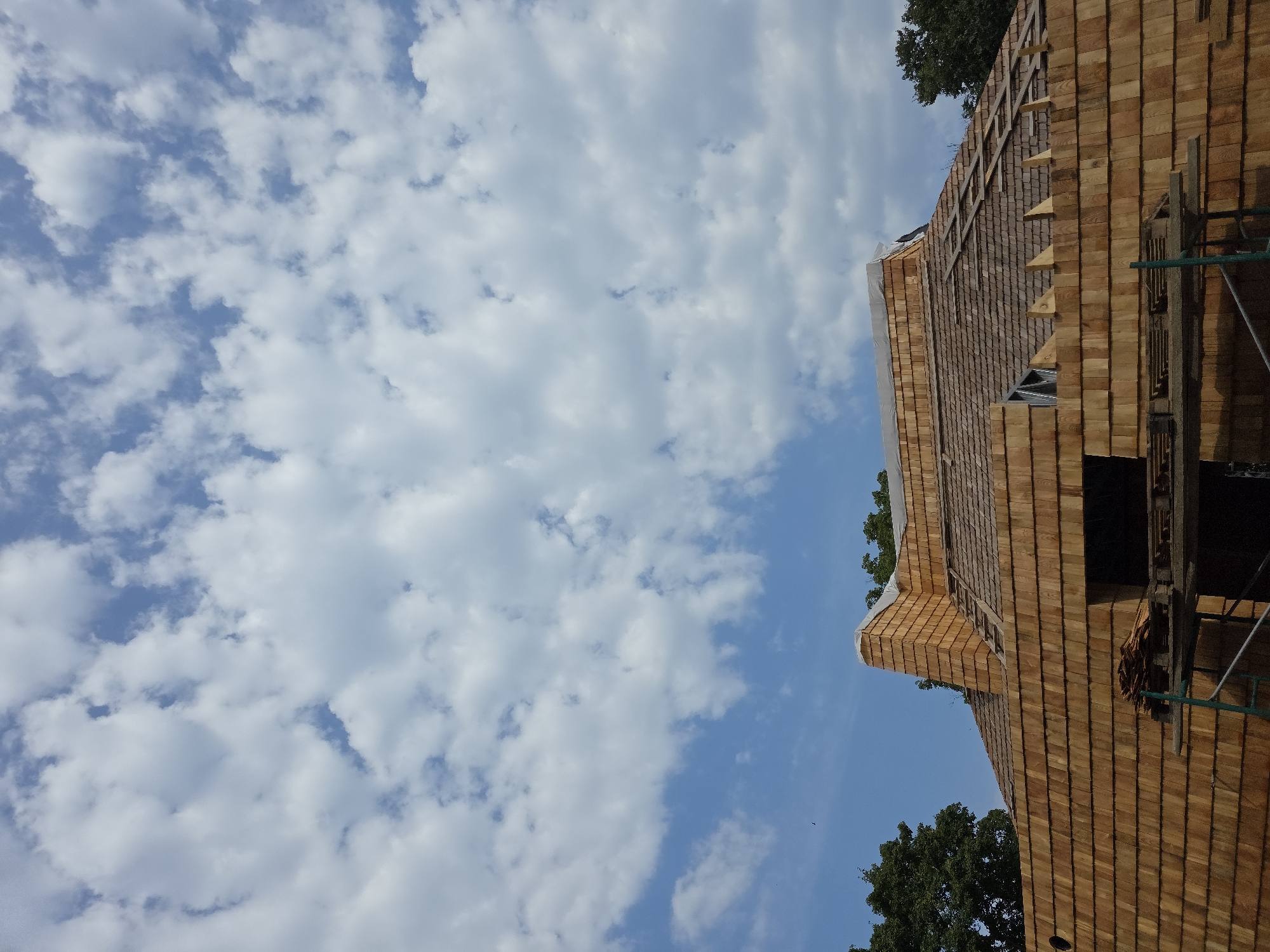 A low-angle exterior shot captures a building under construction, its roof and walls clad in warm-toned wooden shingles. The sky above is a vibrant blue, dotted with fluffy white clouds, suggesting fair weather. The building appears to be a substantial structure, possibly a house or other dwelling, with visible scaffolding and construction materials in the foreground. The surrounding environment includes lush green trees, indicating a natural or semi-rural setting. The image does not contain any people. The specific location in the city of Mamaa'ul, Russia, is not discernible from the visual information provided.