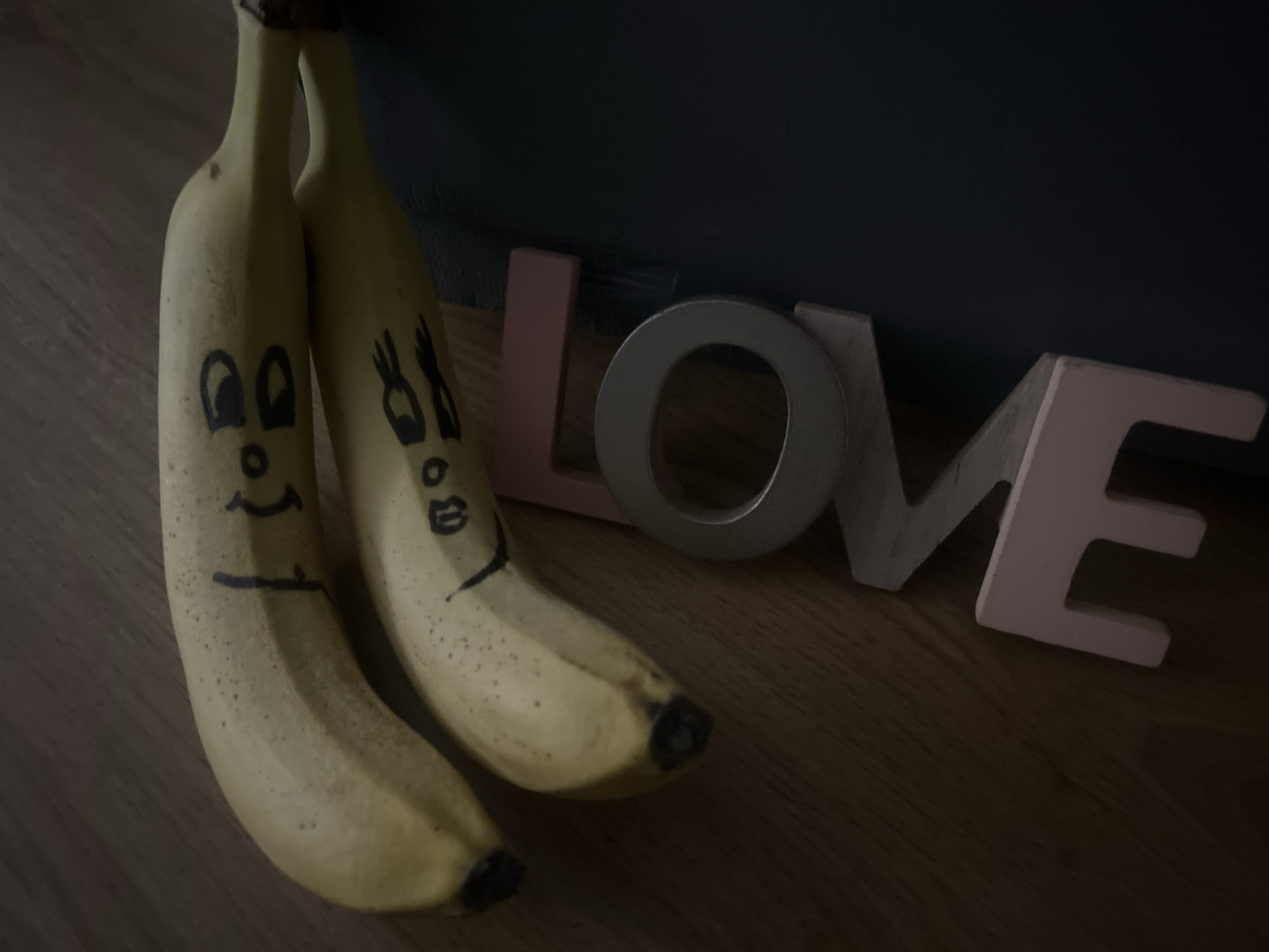 Two bananas are positioned on a wooden surface beside the word "LOVE" spelled out in individual wooden letters of varying colors: pink, silver, and white. Faces have been drawn on the bananas with a black marker. The background is a dark teal color. The location is unspecified, but the setting is indoors.