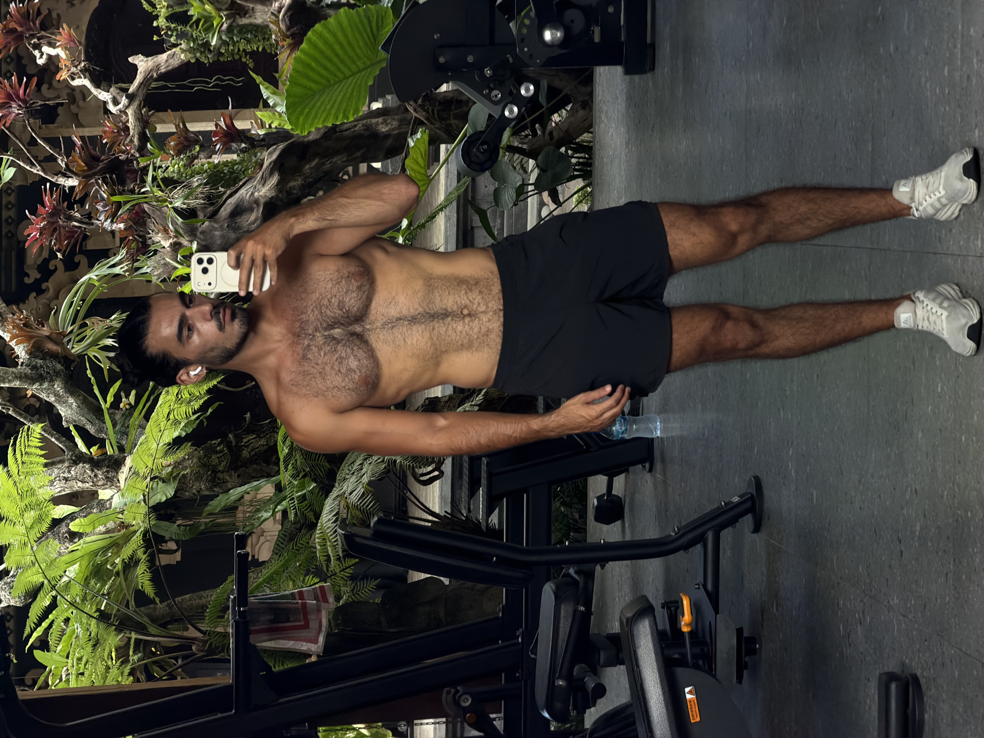 A shirtless, athletic man is posing for a selfie in what appears to be a gym in North Kuta, Indonesia. He is wearing dark shorts and white sneakers. He has dark hair pulled back into a bun and is holding a white smartphone. The background includes gym equipment, plants and trees.