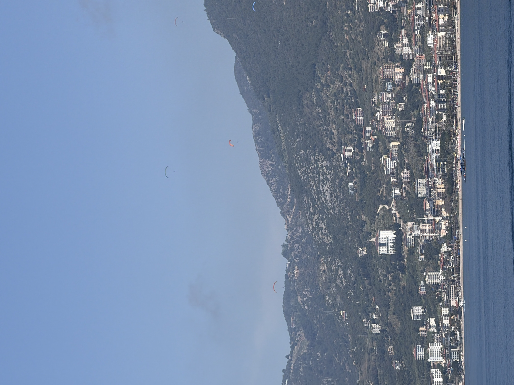 The image captures a panoramic view of a coastal town in Alanya, Turkey, under a clear blue sky. The scene is oriented vertically, with a vast expanse of light blue sky on the left. Several paragliders are visible in the sky: an orange paraglider is somewhat central, a light blue one is higher up, and two off-white ones are further to the left.
Below the sky, a large, steep mountain range covered in dense green vegetation forms a dramatic backdrop. Along the base of the mountains and extending to the right is a bustling coastline with numerous white and light-colored buildings, indicative of a town or city, possibly a resort area. These buildings vary in size and architectural style, suggesting a mix of residential and commercial structures.
To the far right, a calm, dark blue sea stretches into the distance, with a few small boats visible near the shore. The overall impression is one of a vibrant coastal destination popular for activities like paragliding. The bright, clear conditions suggest it is daytime with good weather. There is no visible text in the image.