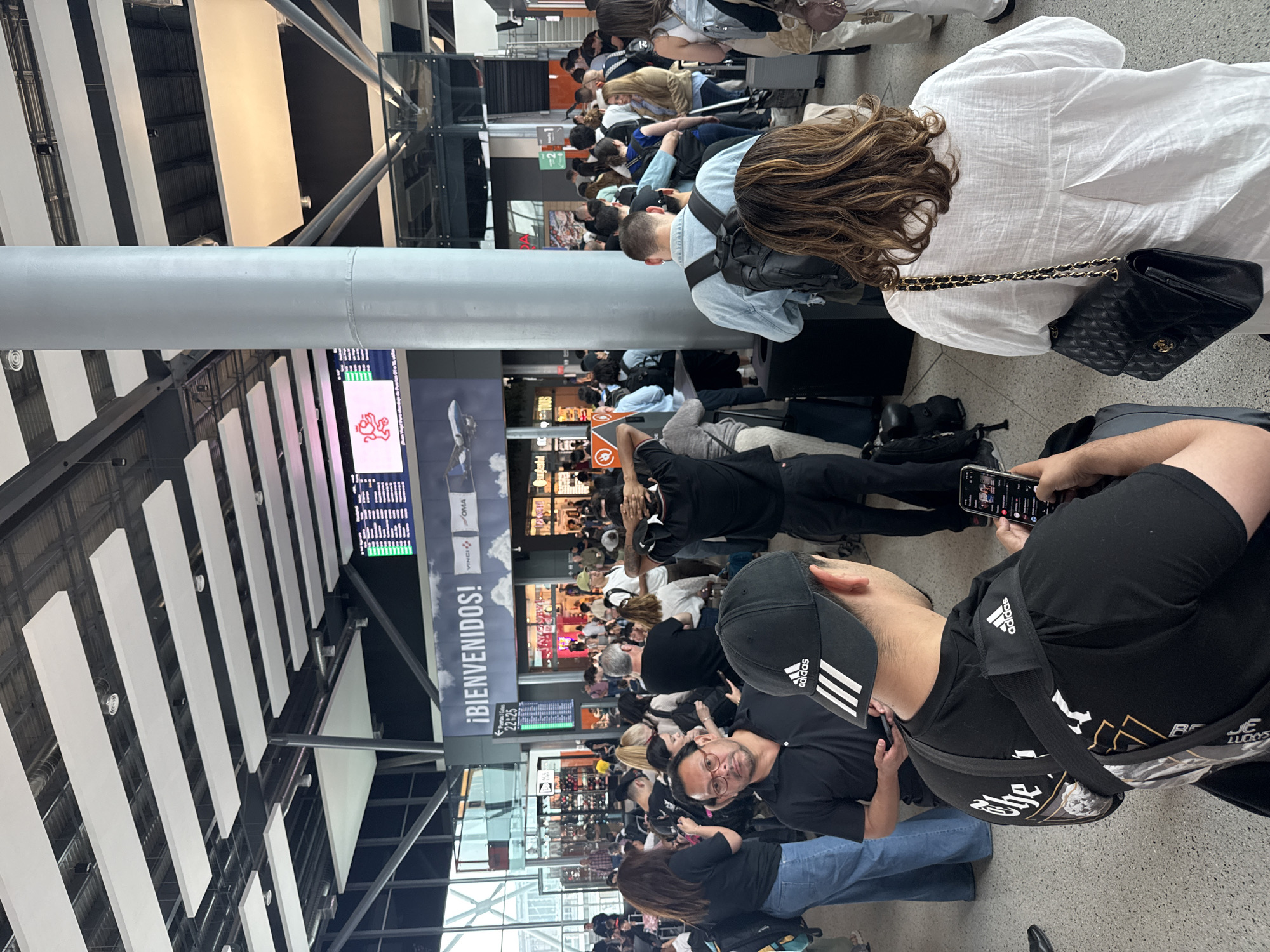 This media file captures a bustling scene inside a modern airport terminal, likely in Apodaca, Mexico, as suggested by the location context and visible cues. The perspective is from behind several individuals in the foreground, looking towards a crowded waiting or check-in area.
Numerous people of various ages are gathered, predominantly standing in lines or congregating. Many are occupied with their mobile phones, either holding them or looking at their screens. Some individuals are seated, while others are standing with luggage, including rolling suitcases and backpacks.
The setting features high ceilings with prominent white rectangular acoustic panels and a grey cylindrical pillar dominating the foreground. Large digital screens display information, some showing what appears to be flight schedules or advertisements. One screen prominently features a red lion-like logo, possibly for an airline like Aeromexico.
Visible text and branding include:
* A large sign proudly displaying "¡BIENVENIDOS!" (Spanish for "WELCOME!").
* On an information screen, "VINCI" and "OMA" (Grupo Aeroportuario del Centro Norte, an airport operator) are visible.
* Another sign indicates gate numbers "22-25".
* The "Adidas" logo is clearly seen on a cap and shirt worn by individuals in the foreground.
* A small sign for "Continental" is also present.
The activity primarily involves waiting, typical for an airport environment. The lighting is bright and artificial, suggesting an indoor setting and no specific time of day like dawn or dusk, but rather general operational hours. The overall atmosphere appears orderly despite the crowd, with people patiently engaged in their personal activities while awaiting their next step in travel.