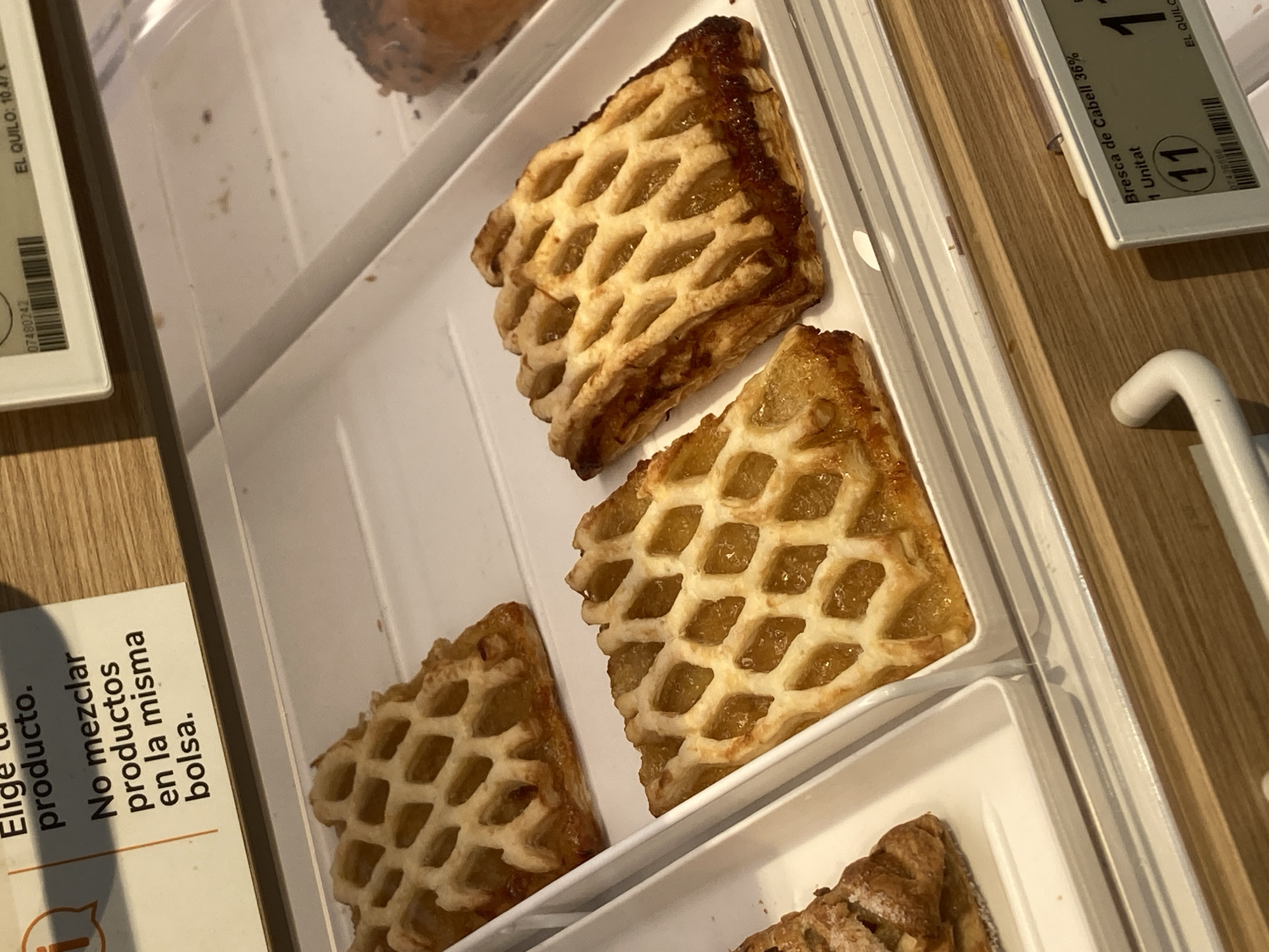 The image shows a close-up, overhead view of several pastries in a display case. The pastries appear to be sweet, with a lattice-like crust filled with a golden, jam-like substance. Some of the pastries have slightly darker, caramelized edges. They are arranged in white rectangular trays.
To the left of the pastries, a white label is partially visible with text in Spanish. Visible words include "Elige tu producto," "No mezclar productos en la misma bolsa," and a barcode.
To the right and top, there are other labels with Spanish text and a numerical indicator. One label reads "Bresca de Cabell 36%," "Unitat," and a number "11" within a circle. Another label has "EL QUILO: 10.47" and a barcode.
The pastries are presented in a setting that suggests a bakery, pastry shop, or a food counter in a market. The lighting is bright, indicating an indoor commercial environment. The overall impression is one of ready-to-eat, appealing baked goods. No people are visible in the image. The presence of Spanish text strongly suggests the location is within Spain, specifically Barcelona as per the context provided.