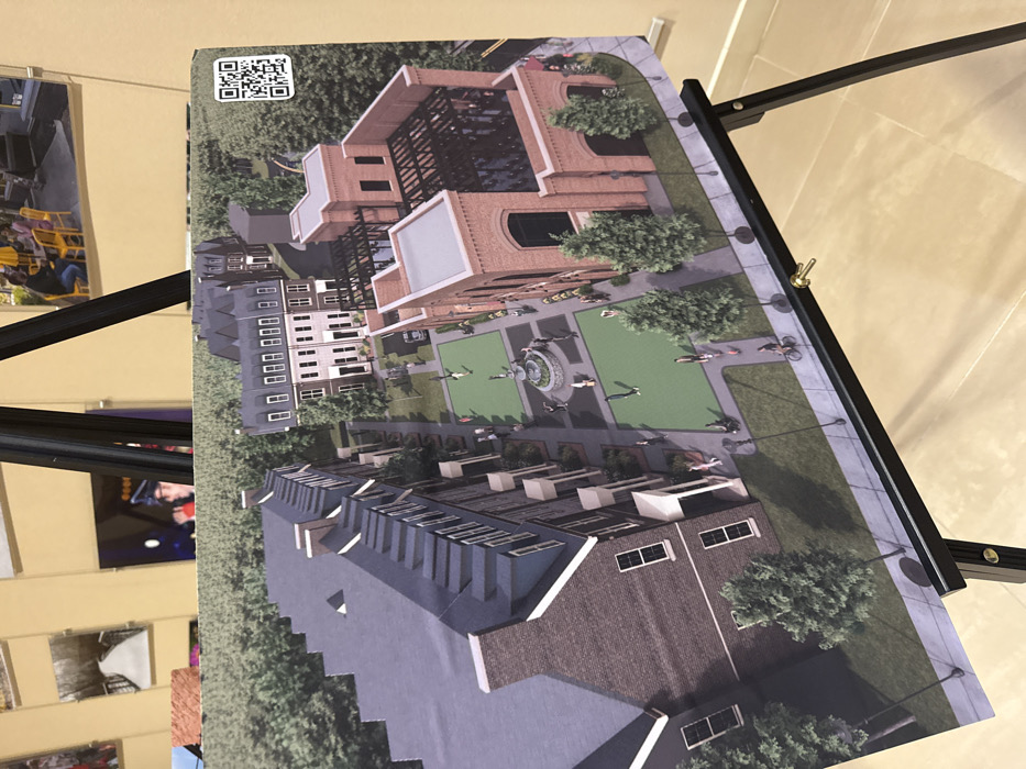 An architectural rendering depicts a planned community. A central green space features a fountain, surrounded by pedestrian paths and people. Townhouses with pitched roofs anchor the bottom of the image. A taller building with a white facade is to the left. A brick building with a modern, open-air top sits at the upper right. A QR code is visible in the upper left corner. The rendering is mounted on a black easel.