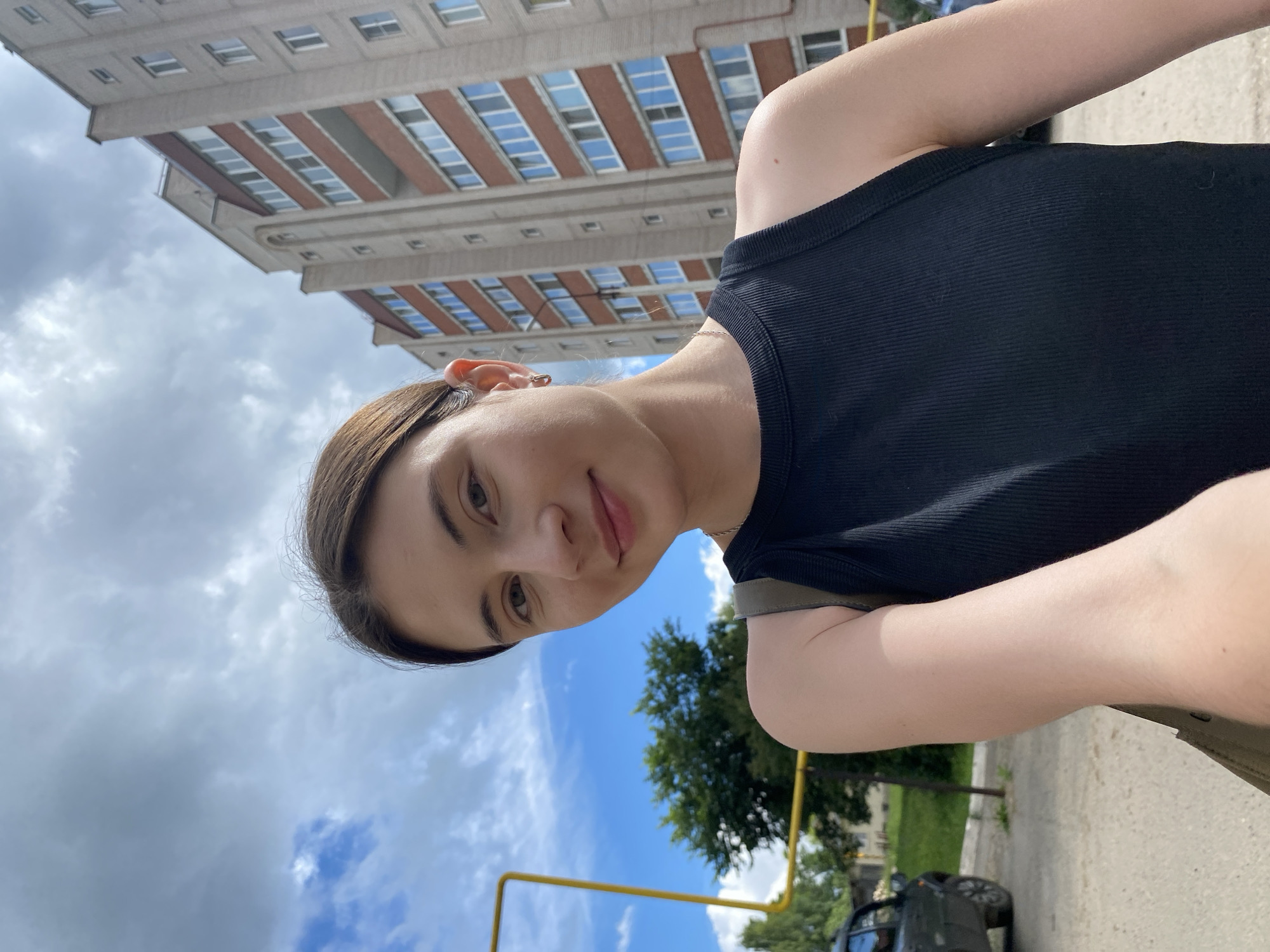 A young woman with brown hair and fair skin is taking a selfie outdoors. She is wearing a black ribbed tank top and a thin necklace. Her left ear is adorned with a small hoop earring. In the background, a tall, multi-story apartment building with red brick accents and numerous windows is visible. To the left of the building, a patch of green trees and a cloudy blue sky can be seen. A yellow pipe structure runs vertically on the left side of the frame, and a dark-colored car is parked in the lower left corner. The scene appears to be a residential area in the city of Votkinsk, Russia.