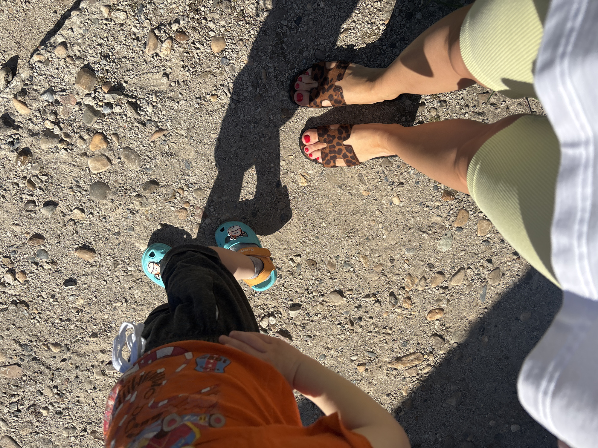 The image shows two adults and a child standing on a gravel surface, possibly a path or road. One adult is wearing leopard print sandals and light green ribbed shorts. The second adult is wearing a white garment. The child is wearing teal clogs, dark shorts, and an orange shirt with cartoon characters. The scene appears to be outdoors in daylight. There is no information in the image that confirms the location as Ферма, Russia.