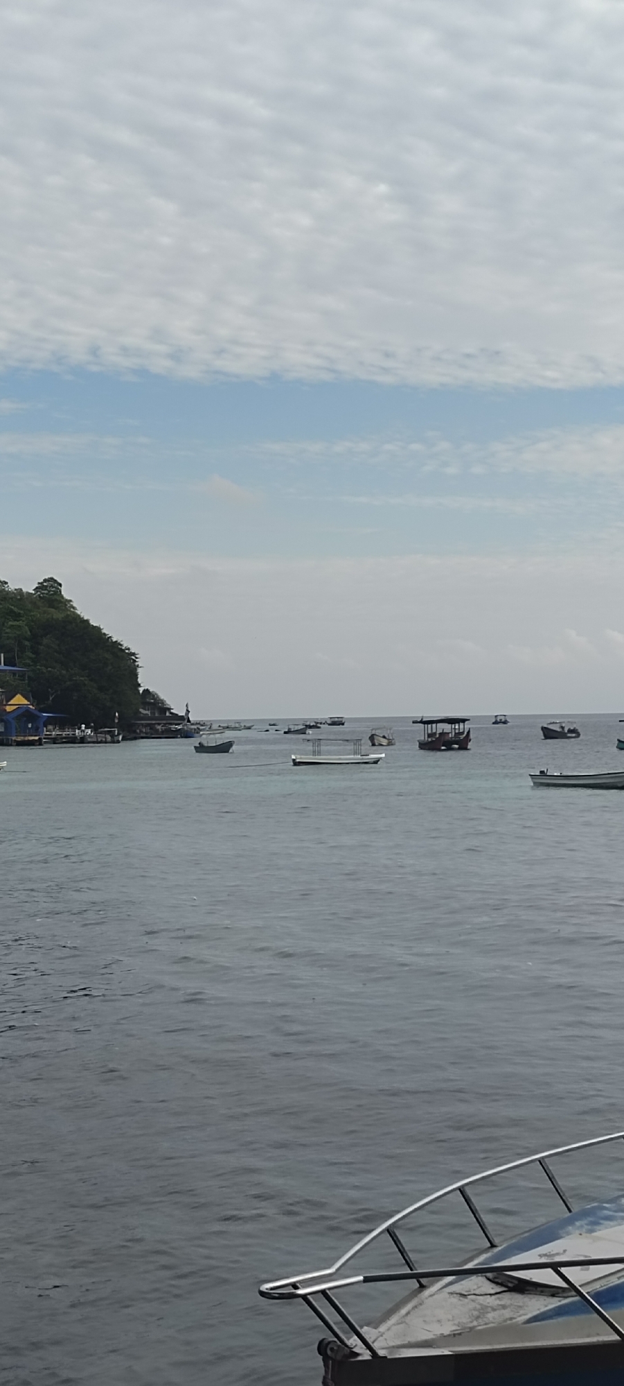 A serene ocean scene unfolds in Sabang, Indonesia, likely near a coastal town as suggested by the presence of buildings and a verdant hillside on the left. The water, a deep grey, is calm with gentle ripples, reflecting the overcast sky above, which is a blend of light blue and scattered, wispy clouds. Numerous boats of various sizes are dispersed across the water. Several are small, wooden vessels, while others appear to be larger, possibly for tourist transport, some with canopies. In the foreground, the metallic railing and part of the hull of a boat are visible, suggesting the vantage point is from another vessel. The overall atmosphere is peaceful and tranquil, indicative of a typical day at sea in this coastal Indonesian locale.