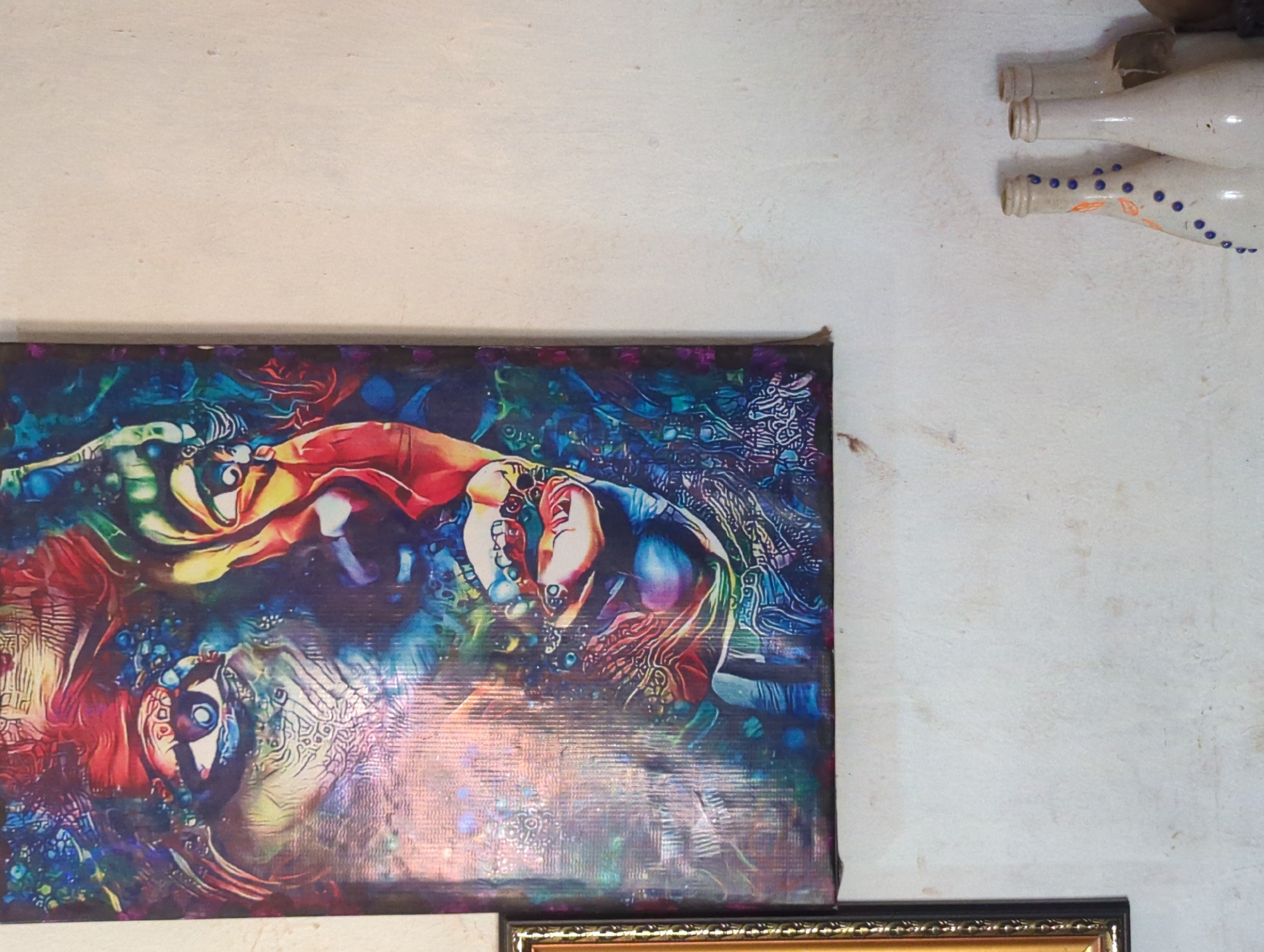 A close-up shot shows a vividly colored, abstract painting hanging on a textured, white wall. The painting features a complex design with human-like figures and faces interwoven with swirls of blue, red, green, and yellow. Below the painting, a portion of a golden-framed piece is visible. In the upper right corner of the image, a decorative display of repurposed glass bottles is attached to the wall. This image was taken in Sabon Tasha, Nigeria.